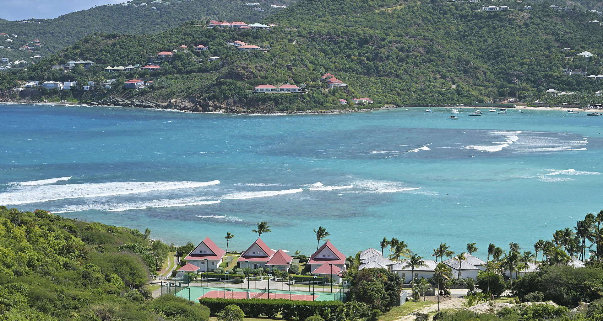 Scenic aerial view of red-roofed villas and tennis court by the turquoise bay, surrounded by lush green hills in St Barts