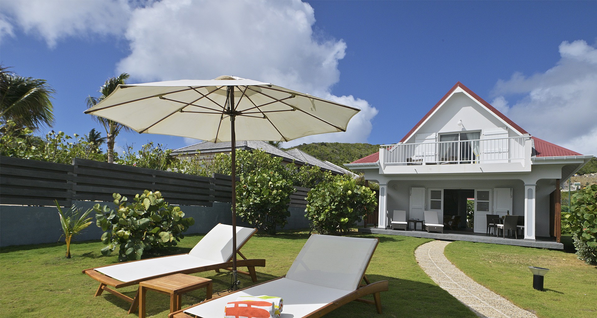 Garden with sun loungers and large umbrella facing a white villa with red roof in St Barts vacation rental.