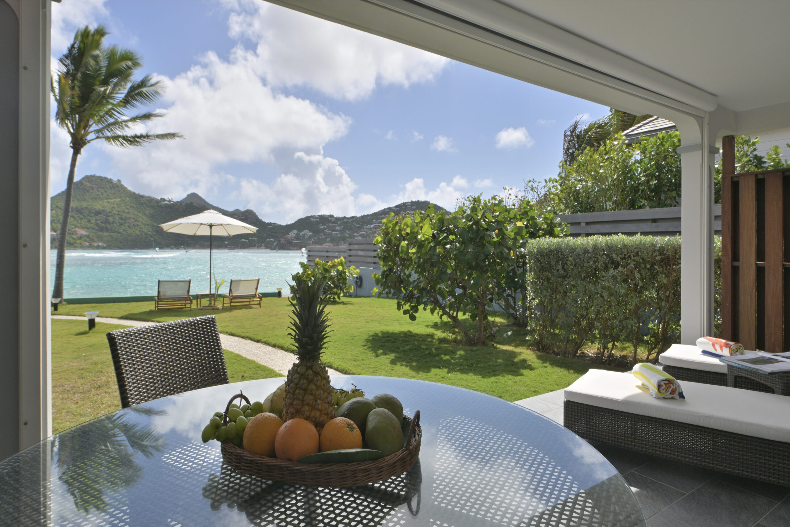 Covered terrace with fruit bowl on glass table overlooking garden, palm trees, and Caribbean Sea at a St Barts villa rental.