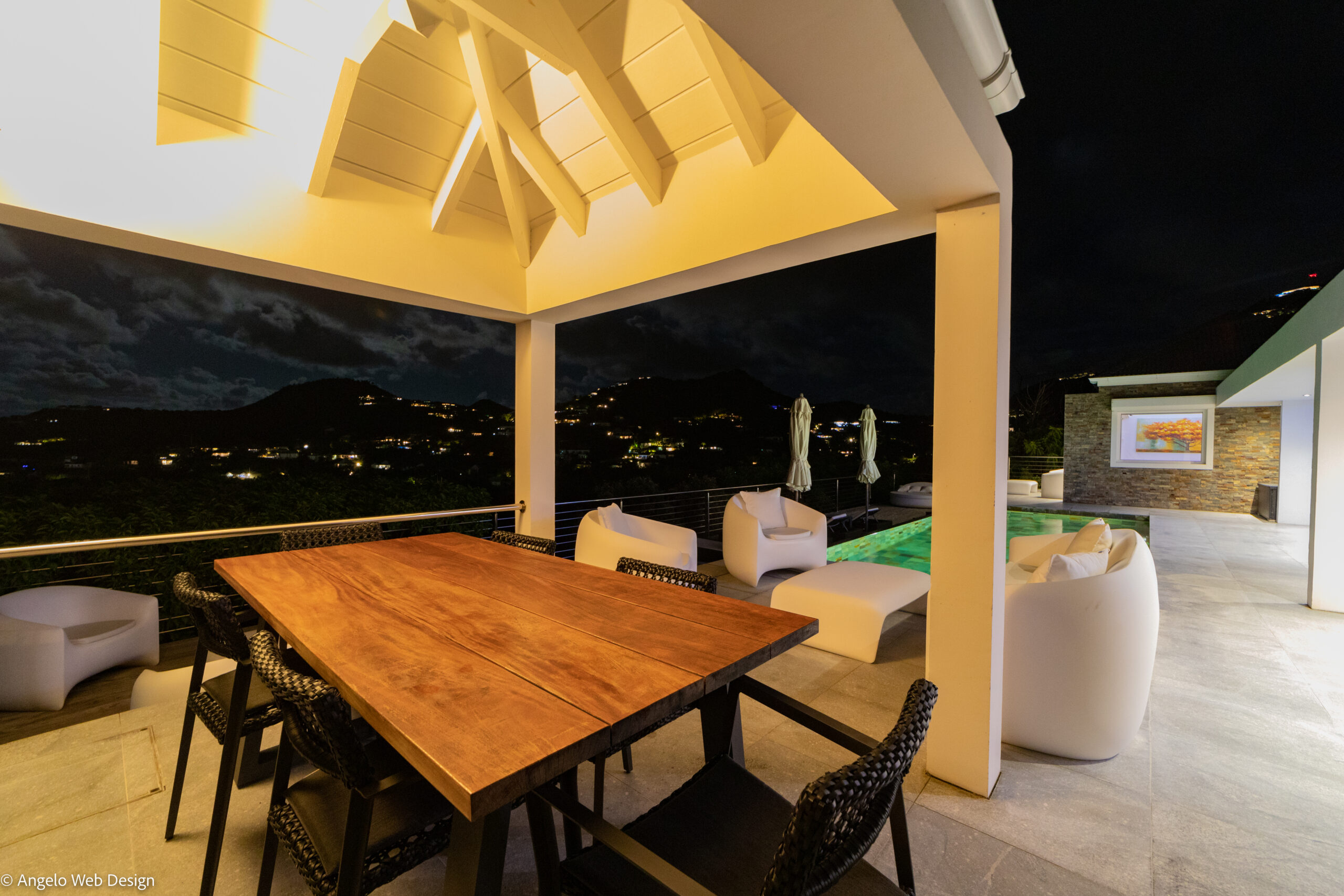 Covered outdoor dining terrace with warm lighting, lounge seating, and poolside views at night in a St Barts villa rental