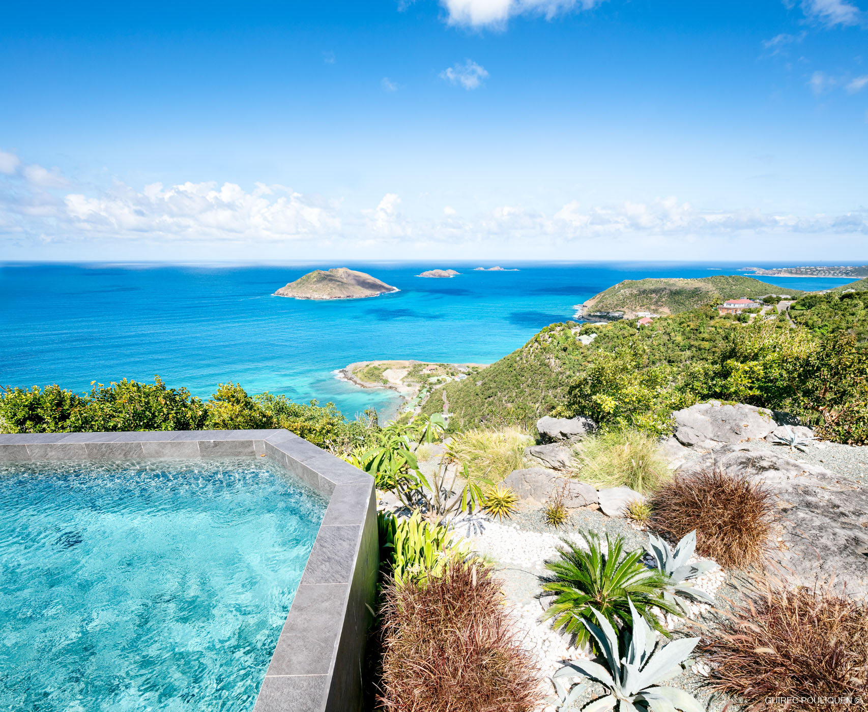 The corner of a swimming pool at Villa Byzance, with the water reflecting the blue sky. The background shows a panoramic view of the sparkling turquoise ocean, a small bay, and distant green islands.