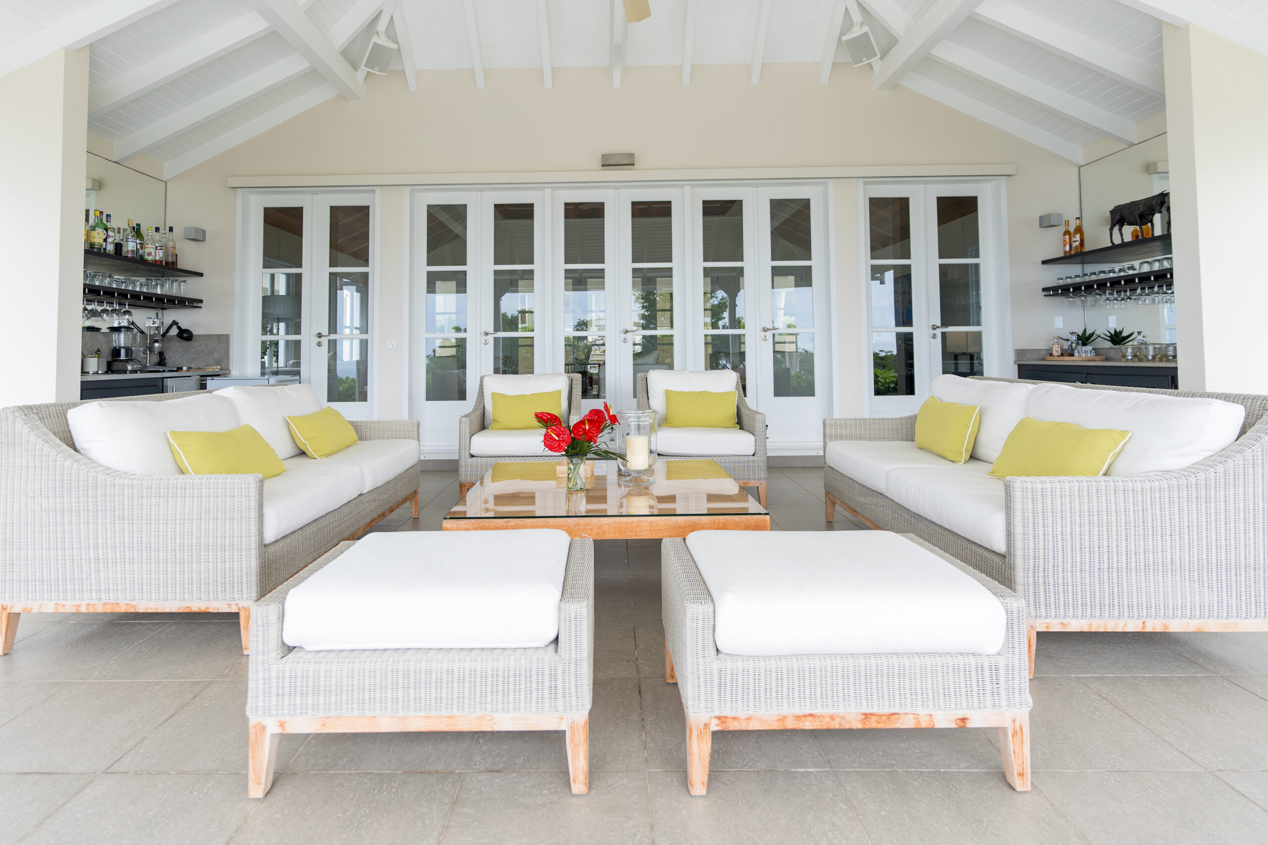 A symmetrically arranged outdoor seating area on a covered patio with two wicker sofas and armchairs with white and lime green cushions, a wooden coffee table, and built-in bar shelves on the side walls.