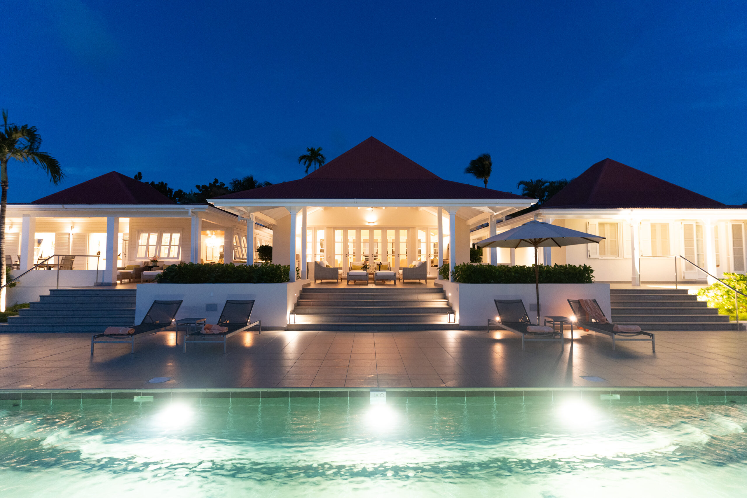 A wide exterior shot of the villa at night, with the main house and pool illuminated by soft lights, and elegant stone stairs leading to the entrance.