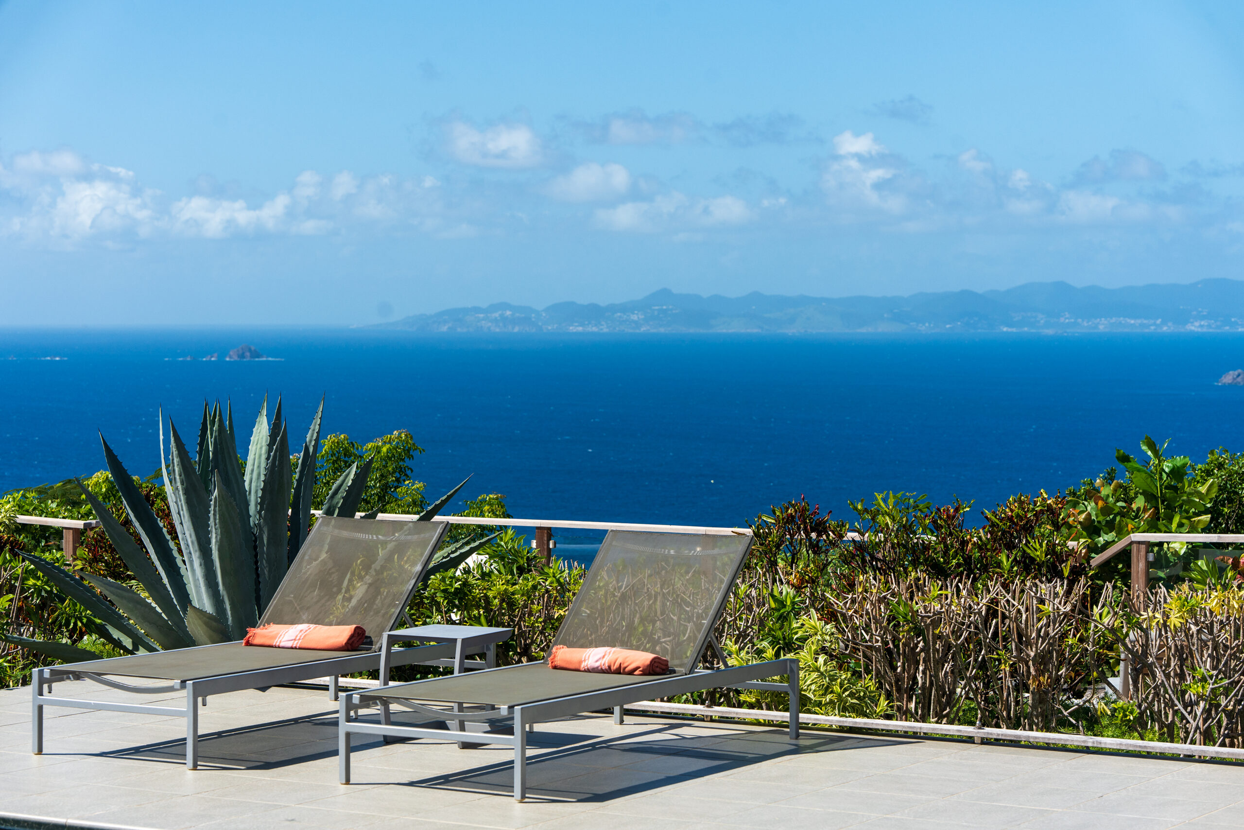 two modern sunbeds on a stone deck, with lush tropical plants and a stunning panoramic ocean view in the background.
