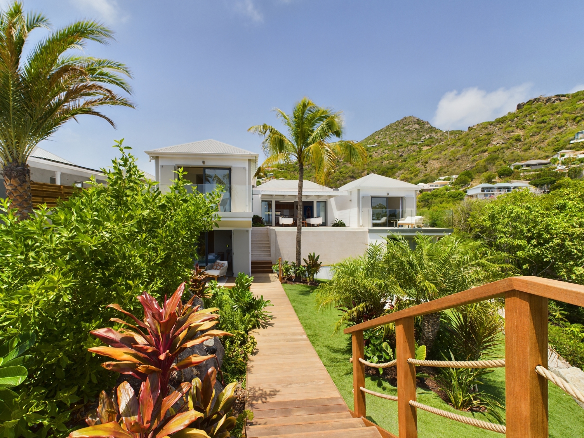 Modern villa in St Barts surrounded by tropical gardens with wooden pathway and mountain backdrop.