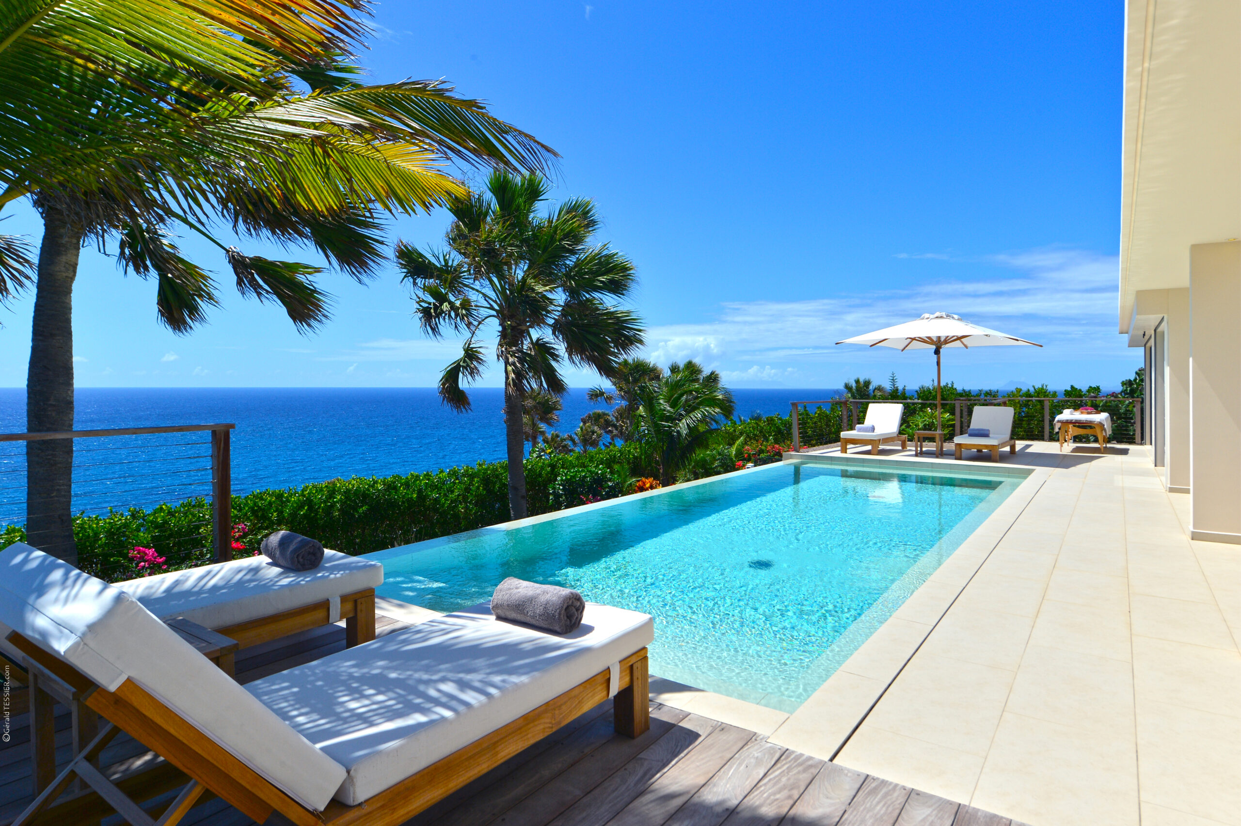 Sunlit infinity pool with white loungers and umbrellas overlooking the Caribbean Sea, framed by swaying palm trees in St Barts