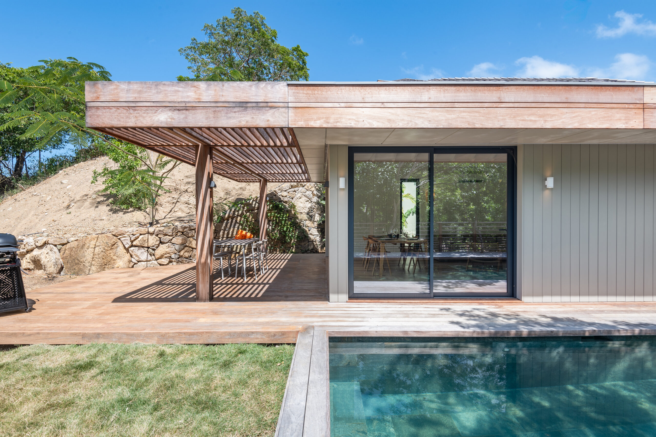 Modern St Barts villa with shaded wooden terrace, sliding glass doors, and poolside deck set against natural rock and greenery.
