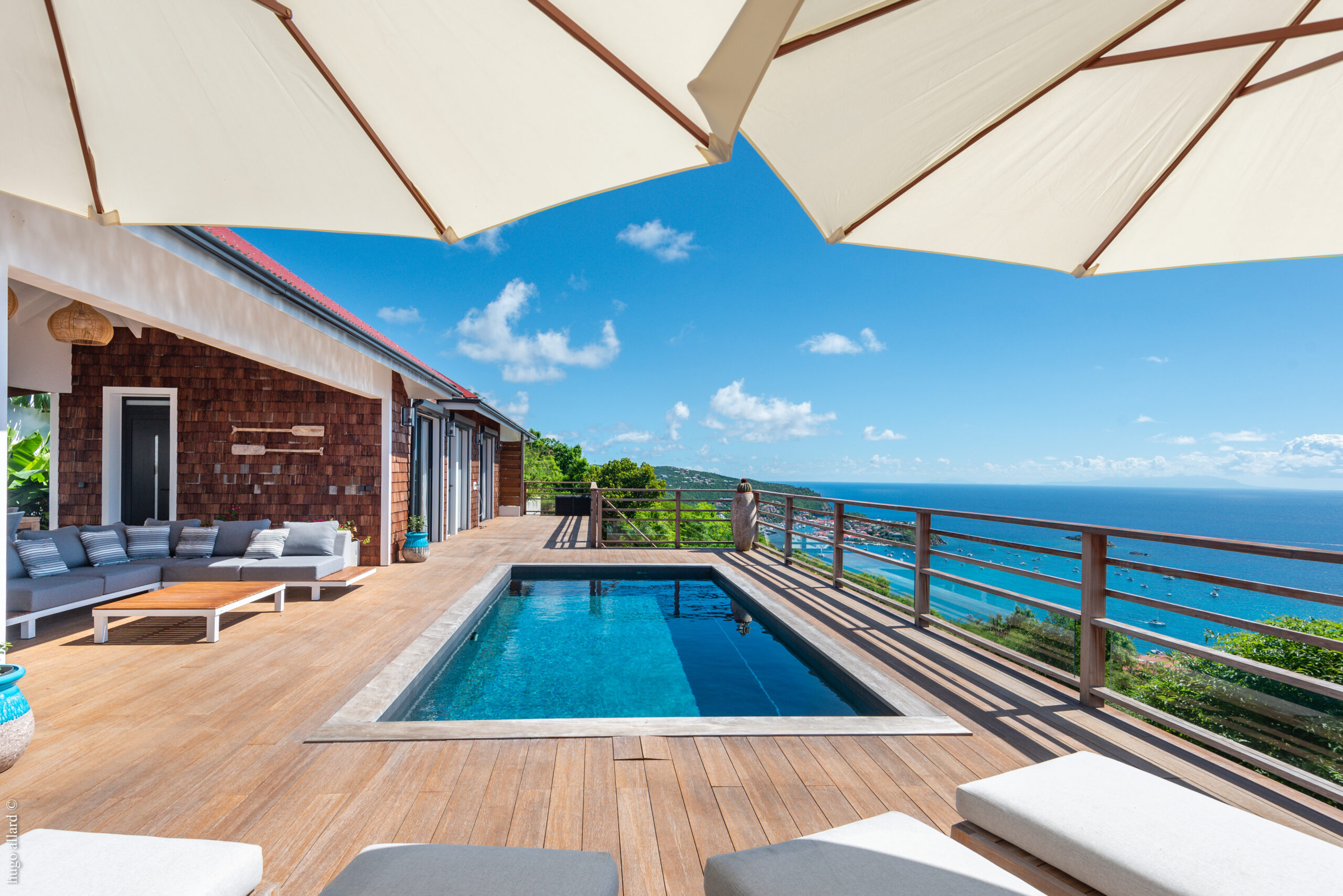 a wooden pool terrace with a small swimming pool, comfortable sun loungers and a modern outdoor sofa set, overlooking a lush hillside and the ocean