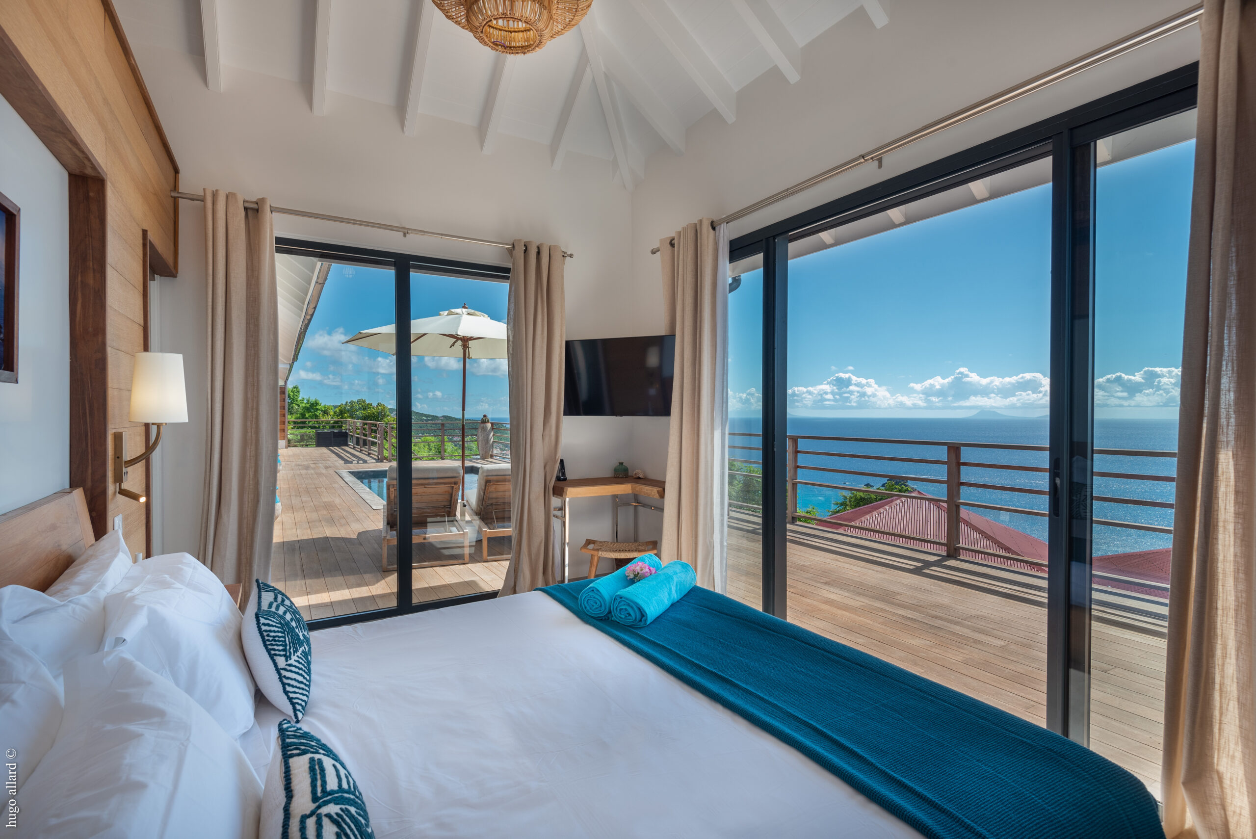 A bright bedroom with a bed with white sheets and a blue throw blanket, a TV on the wall, and large sliding glass doors leading to a wooden deck with a stunning ocean view.