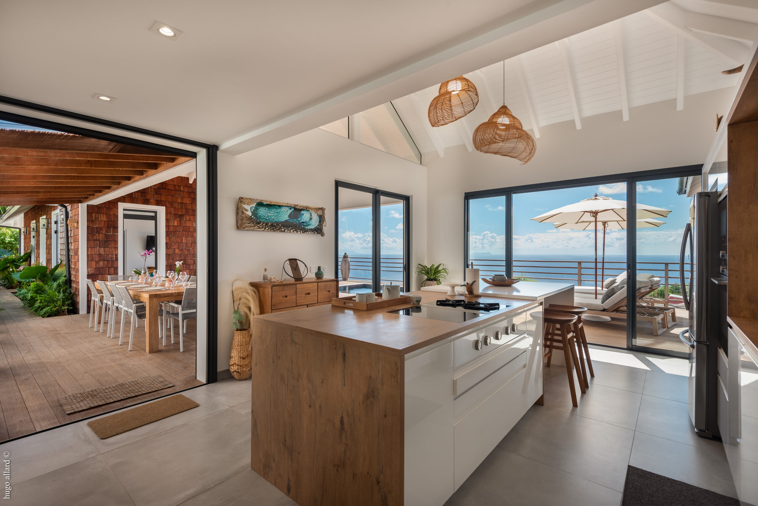 A spacious kitchen with a modern island and stools, sliding doors with an ocean view, and another set of doors leading to a covered outdoor dining area.