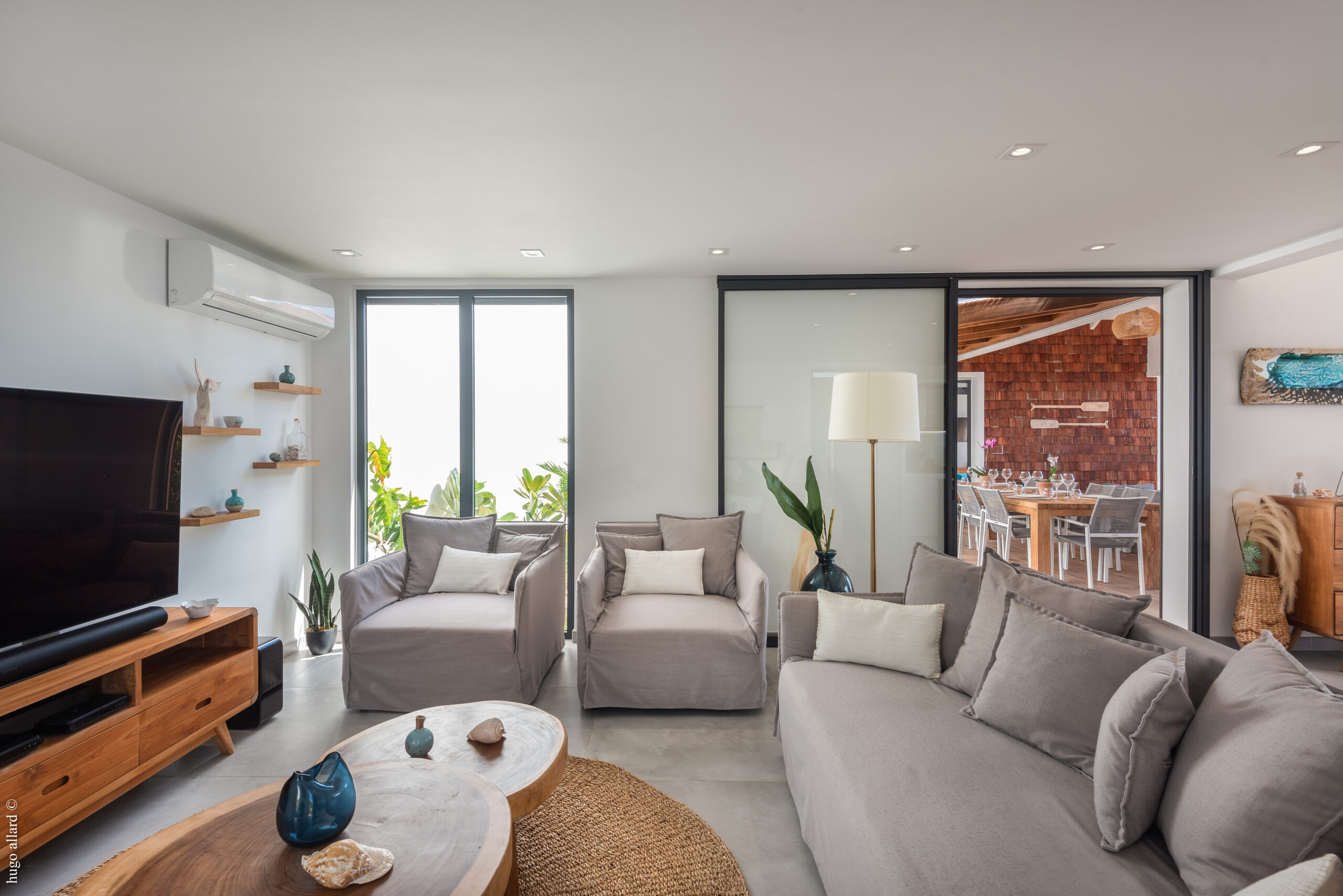 A spacious living room with a gray sofa, two matching armchairs, wooden coffee tables, and a sliding door that reveals a view of an outdoor dining area.
