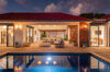 an outdoor terrace at night with a swimming pool and glowing lights, showing a cozy outdoor living room and a modern kitchen area inside the villa