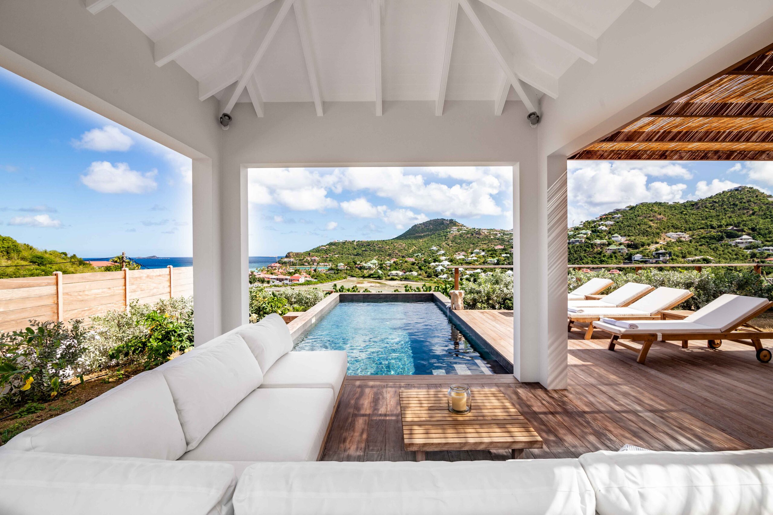 Covered terrace with white sofa and pool deck overlooking green hills and ocean at a luxury St Barts villa rental.
