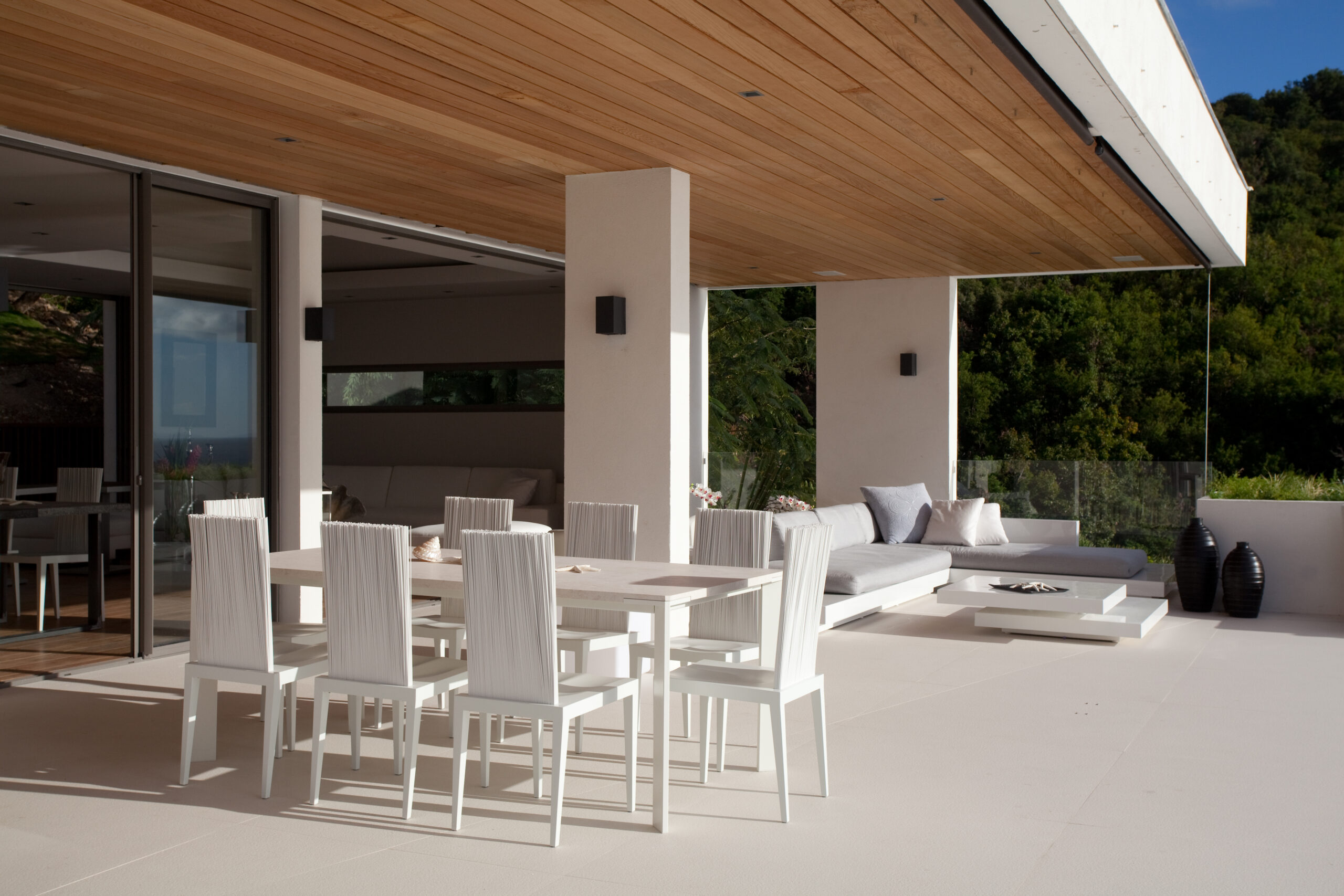 Contemporary outdoor dining area with sleek white chairs, cushioned lounge seating, and a wood-paneled ceiling opening to greenery.