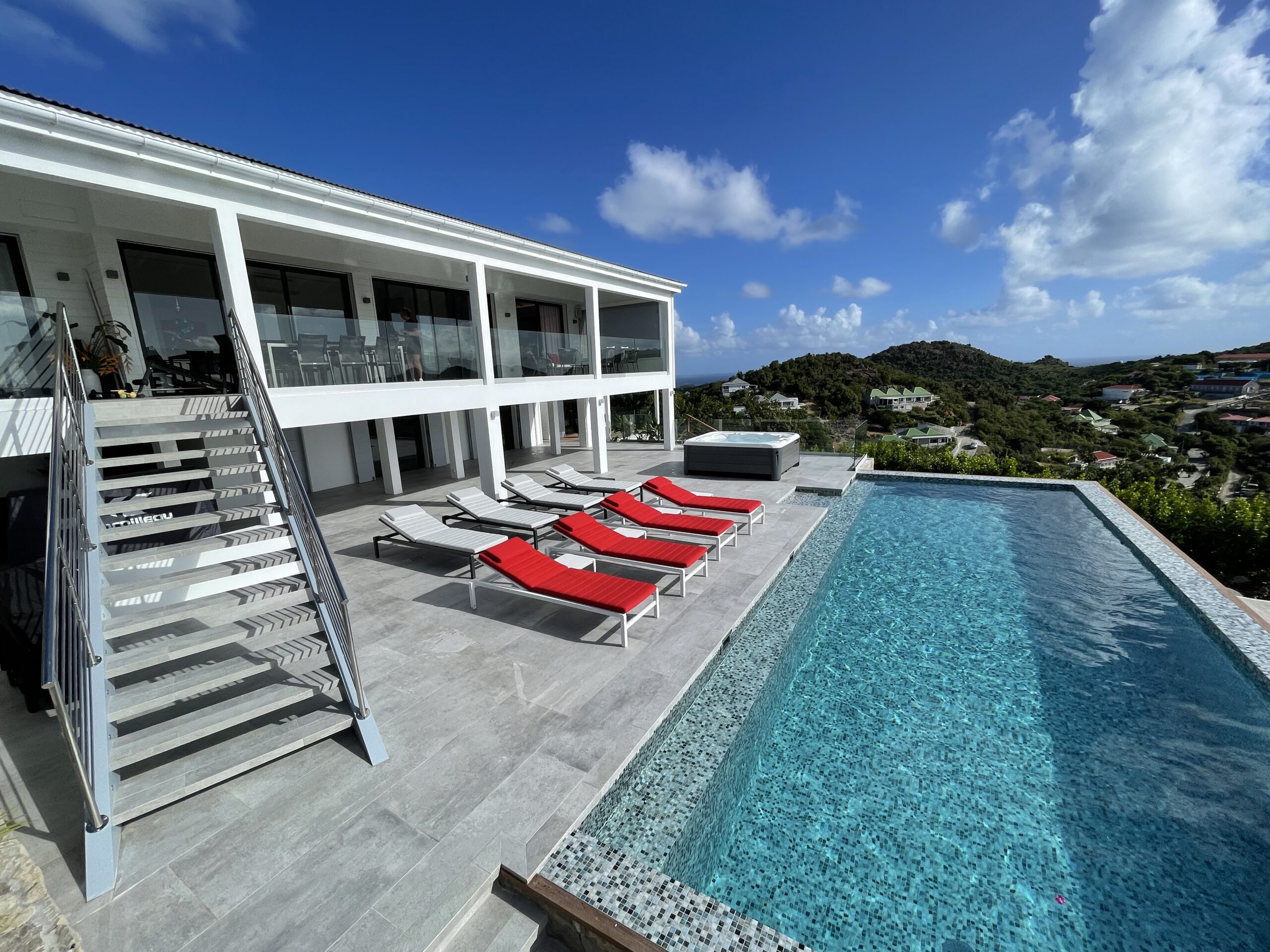 A large outdoor terrace featuring a long infinity pool, a row of red sun loungers, and a modern set of stairs leading to the upper floor of the villa.