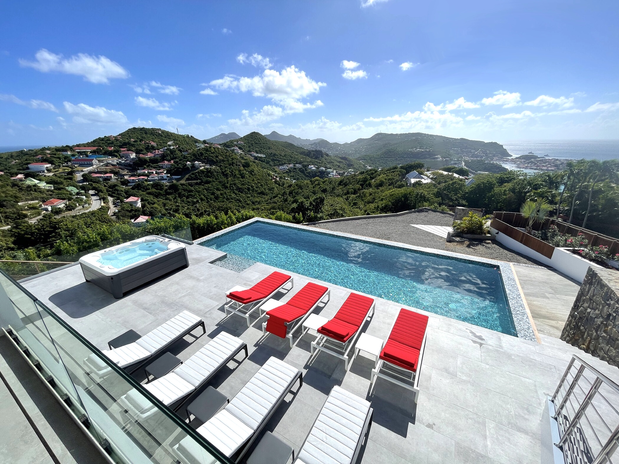 A spacious outdoor terrace with a long infinity pool, a hot tub, and several sun loungers, set against a stunning panoramic backdrop of rolling green hills and the ocean.