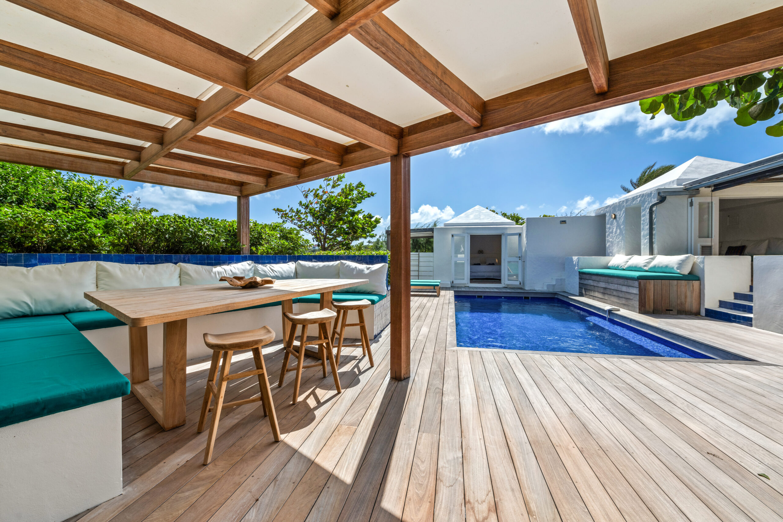 Covered outdoor dining area with wooden pergola, turquoise bench seating, and a view of a private pool surrounded by sun loungers at a luxury St Barts villa rental.
