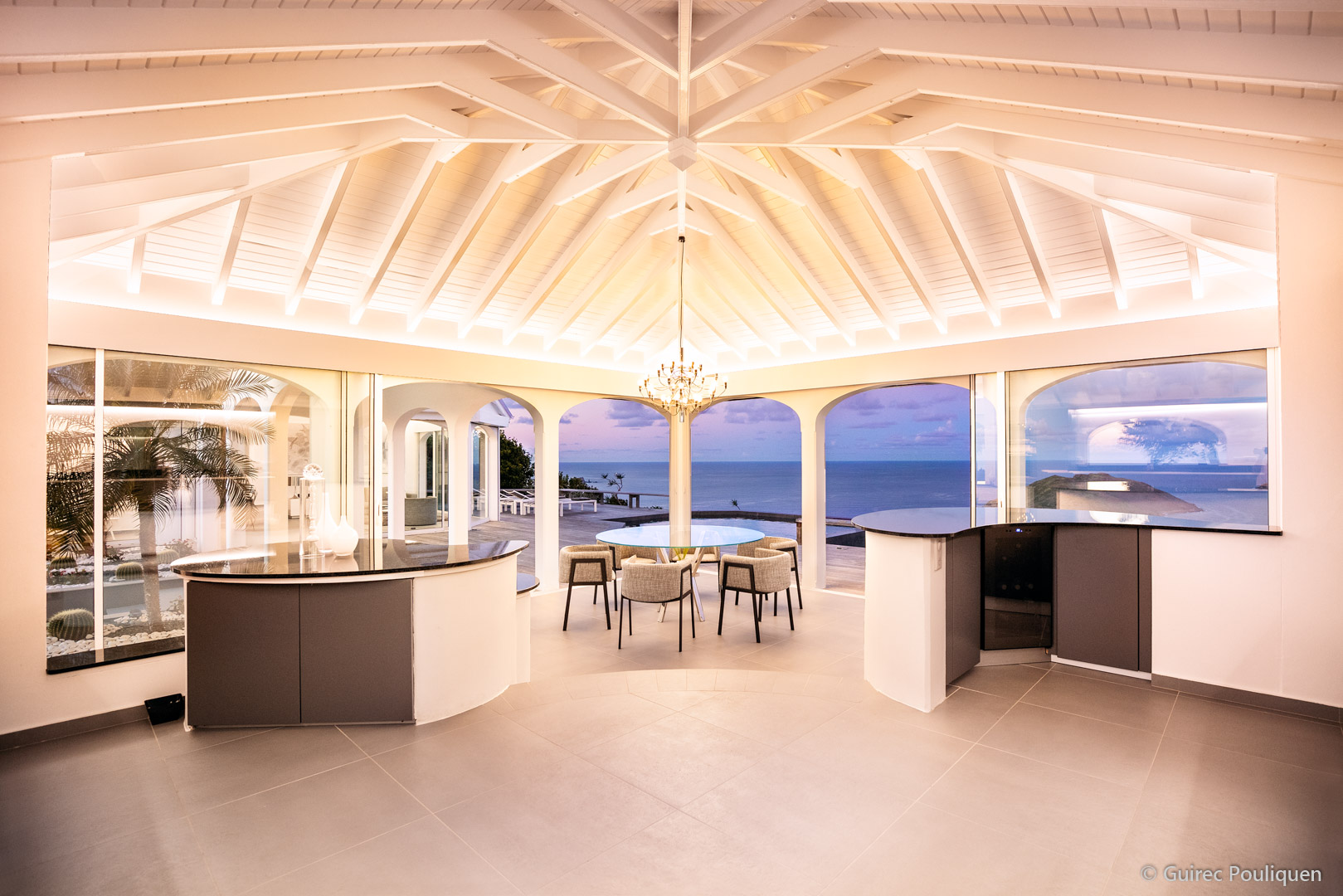 An open-plan kitchen and dining space with vaulted ceilings, a central dining table, and a view of the ocean as the sun sets.