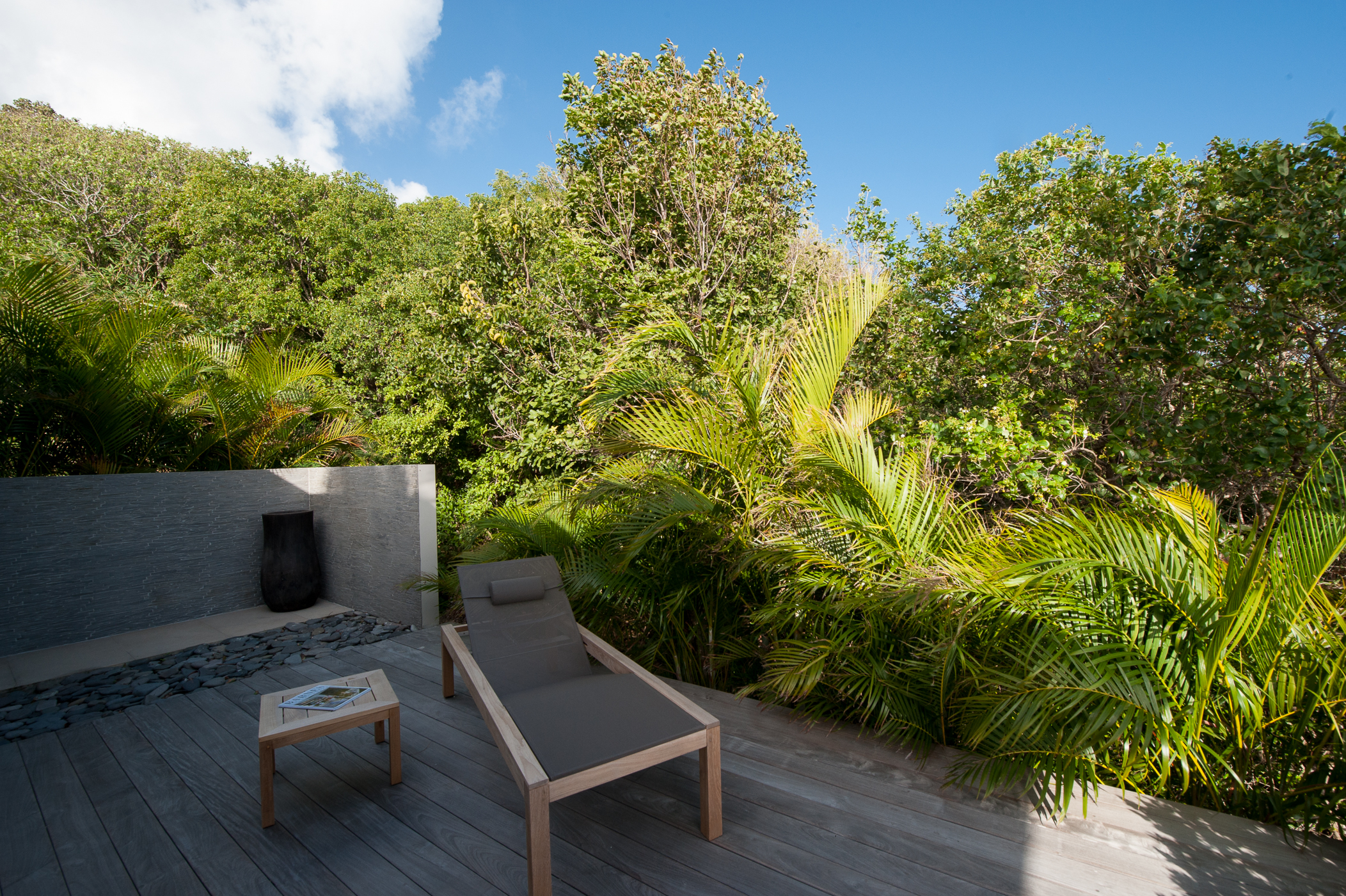 a private wooden terrace with a lounge chair and a small side table, surrounded by lush tropical plants and greenery
