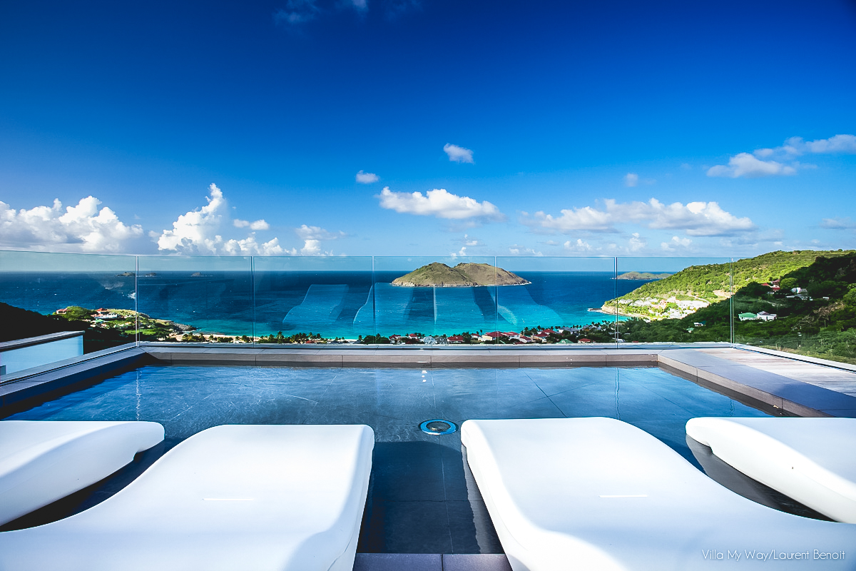 An open-concept living room with a large gray sofa and a unique, tiered coffee table. The view through the open sliding doors shows a wooden deck, a pool, and the ocean and hills at twilight.