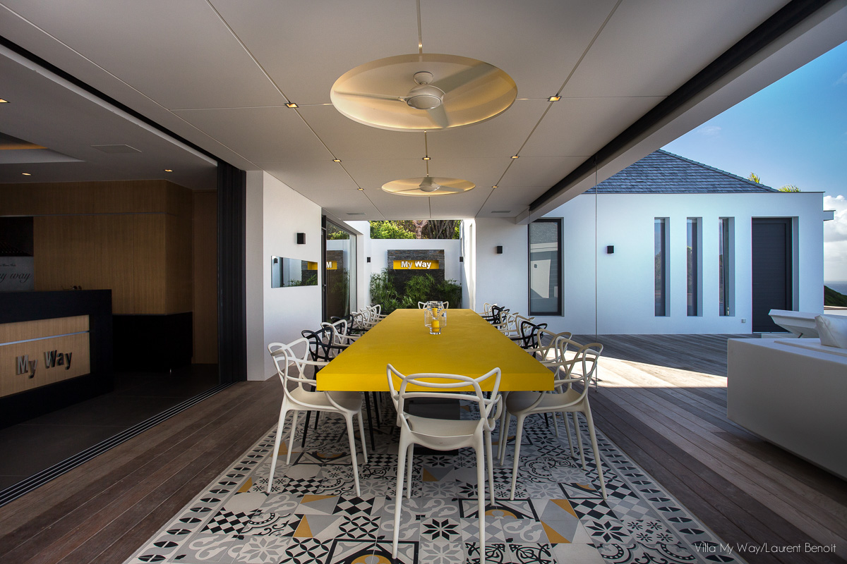 a long shot of the outdoor dining area with a long yellow table on a patterned rug, looking toward the villa's architecture and lush greenery.