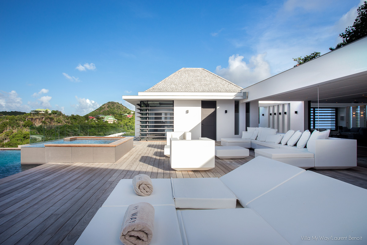 The spacious wooden deck of a modern villa with an infinity pool in the background and a sunken jacuzzi in the mid-ground. White sun loungers and outdoor sofas are arranged to face the ocean and hillside view.