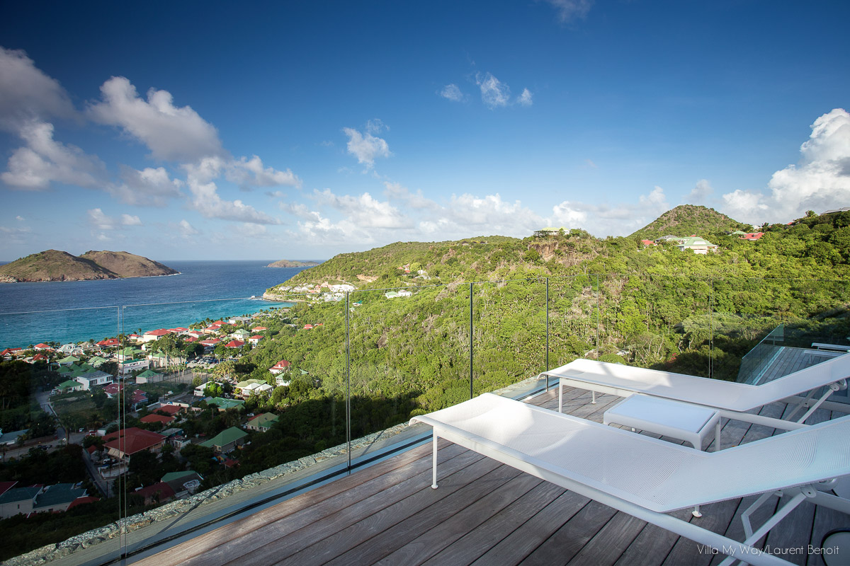An expansive wooden deck with a glass railing, showcasing a view of a lush green hillside, a town with red rooftops, and the bright blue ocean with a distant island. White sun loungers are placed on the deck.