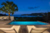 Evening view of a St Barts villa pool with lounge chairs, palm tree, and ocean bay under twilight sky.