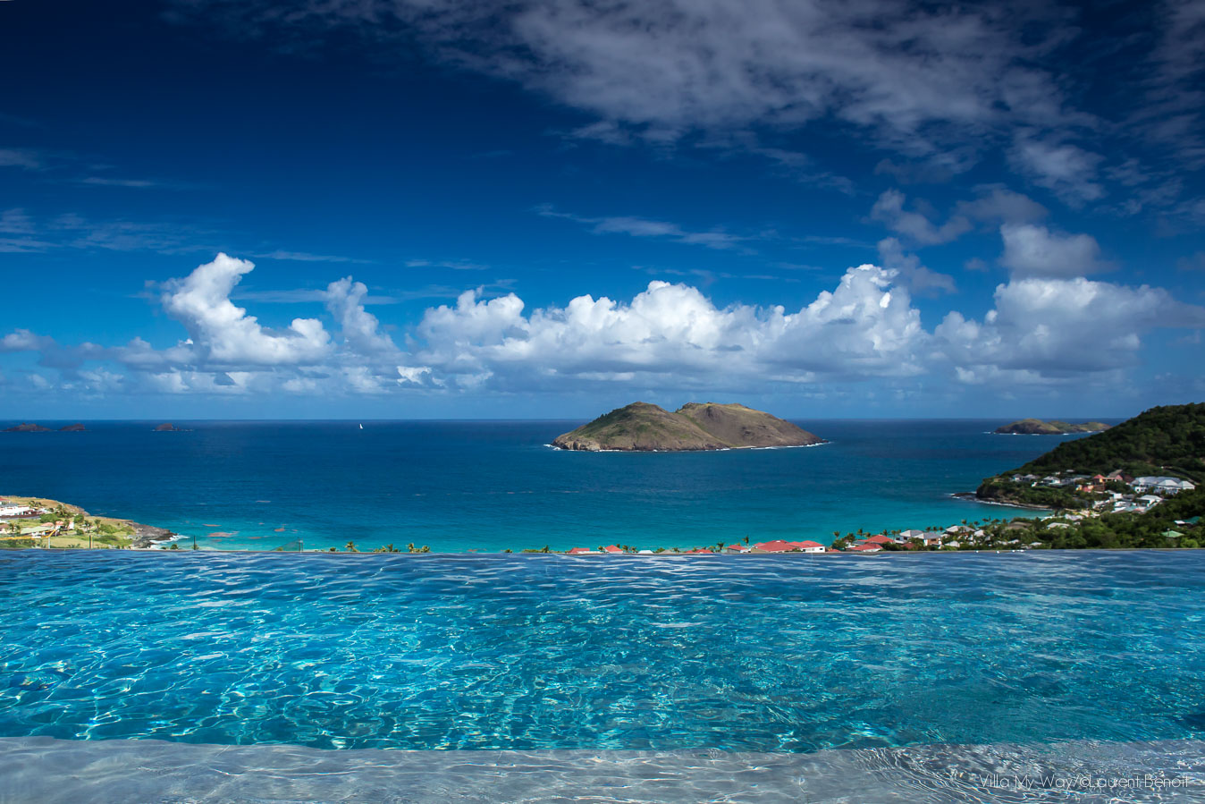 A luxurious infinity pool with white sun loungers on a deck, looking out to a panoramic view of the sparkling turquoise ocean, a distant island, and the St. Barts coastline under a bright blue sky.