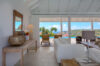 Airy living space with white sofas, wooden accents, and poolside views opening to hillsides in a St Barts villa rental.