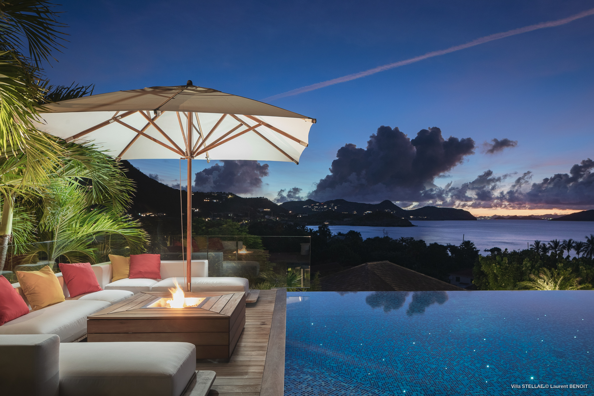Evening poolside scene at Villa Stellae in St Barts with fire pit, cushioned seating, and umbrella overlooking a glowing bay at twilight.