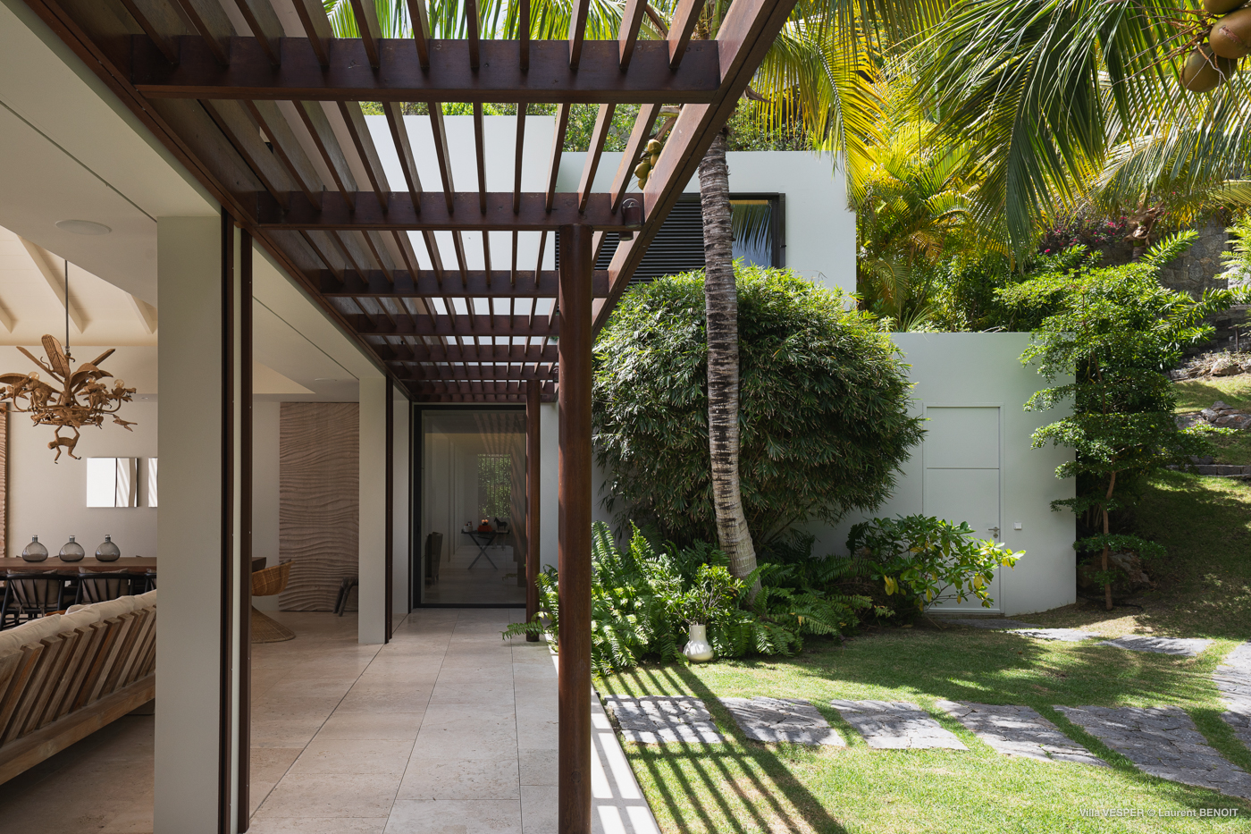 Shaded walkway with wooden pergola leading from an airy living space to lush tropical gardens at a St Barts villa.