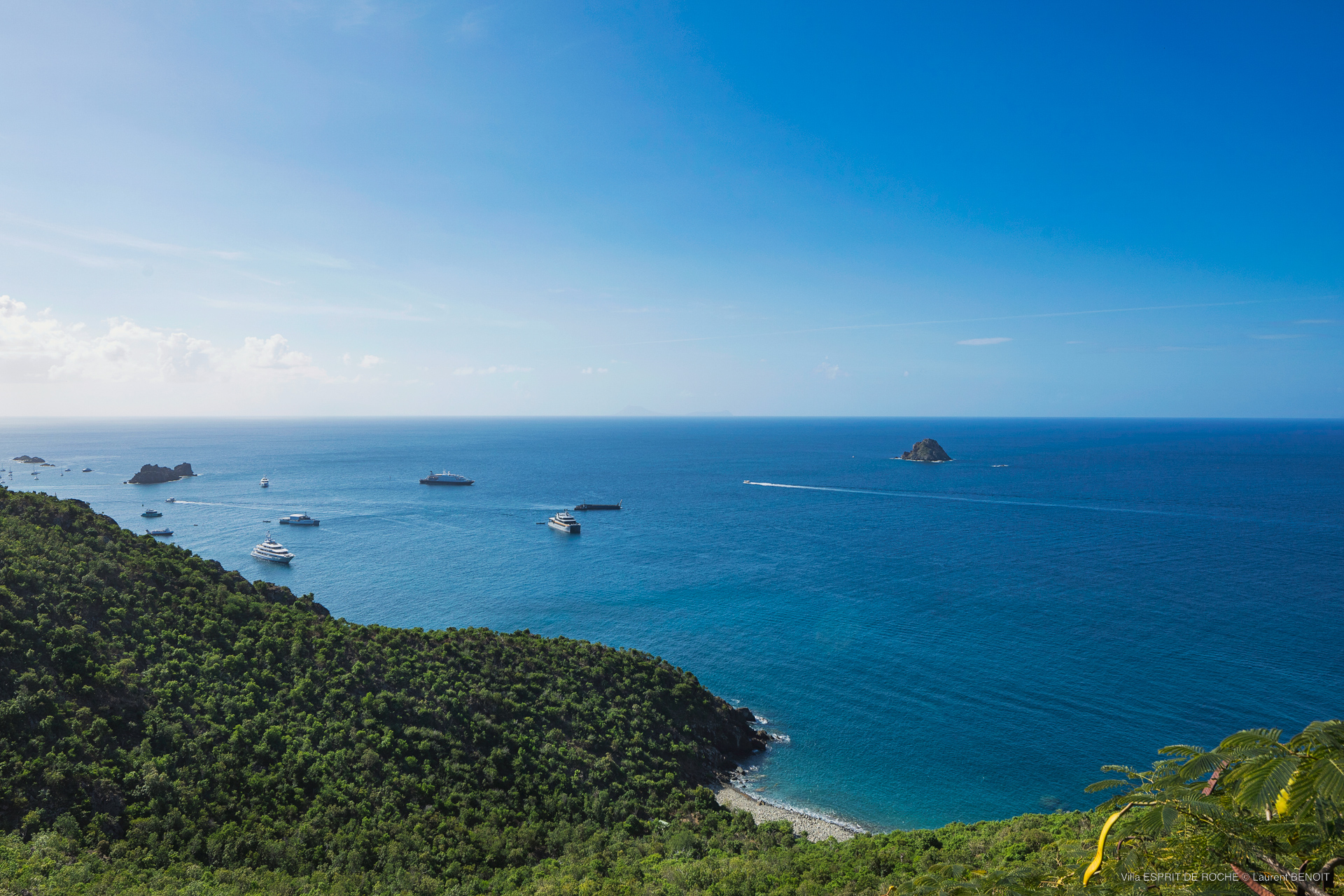 a wide, panoramic shot of the calm blue ocean with several boats in the bay, a small island, and green tropical hills