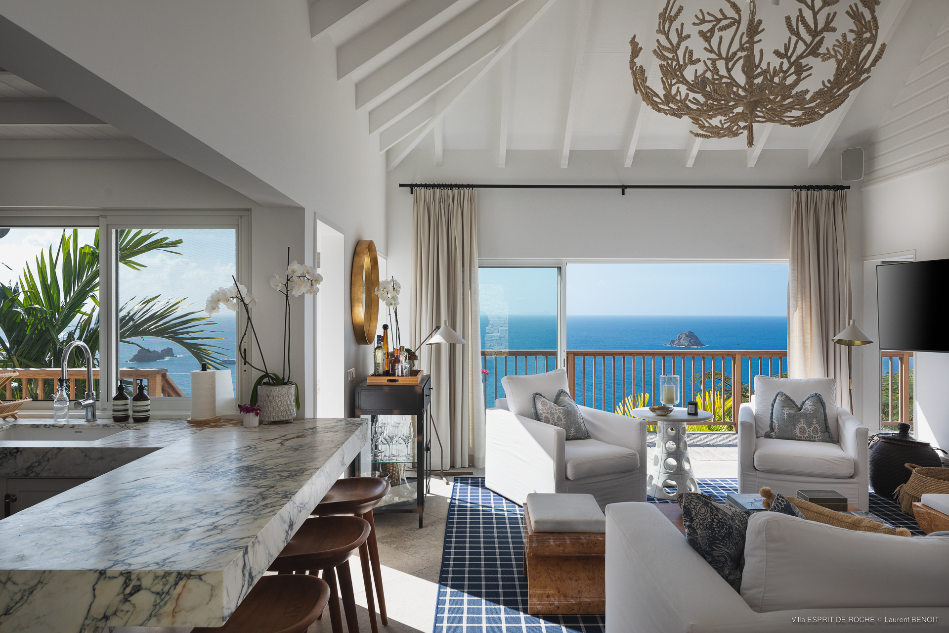 a modern open-concept living area with white couches and chairs, a marble countertop breakfast bar with stools, and a glass sliding door overlooking the ocean