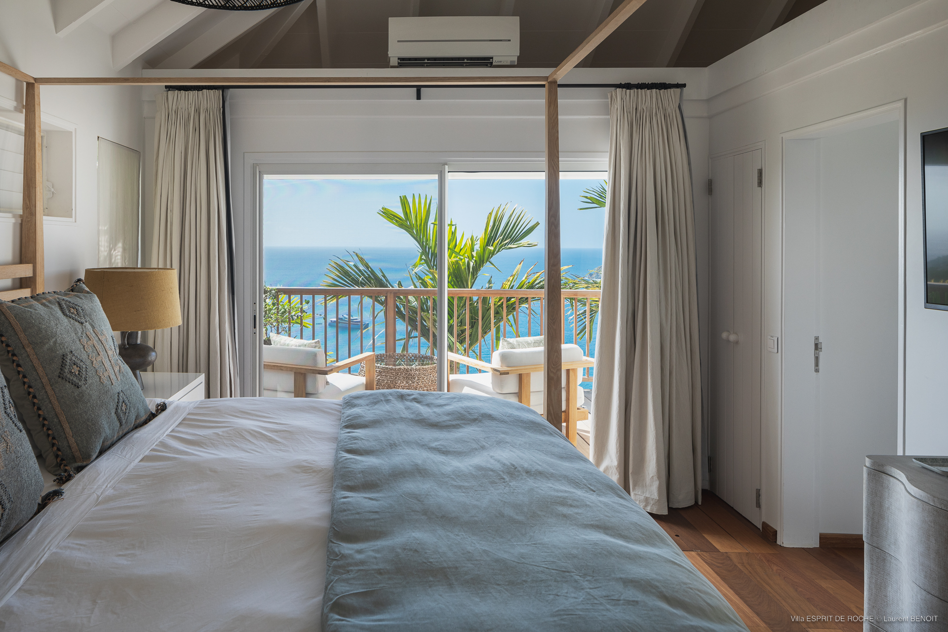 a bright bedroom with a four-poster bed, a light blue blanket, and glass sliding doors that open to a private balcony with an incredible ocean view