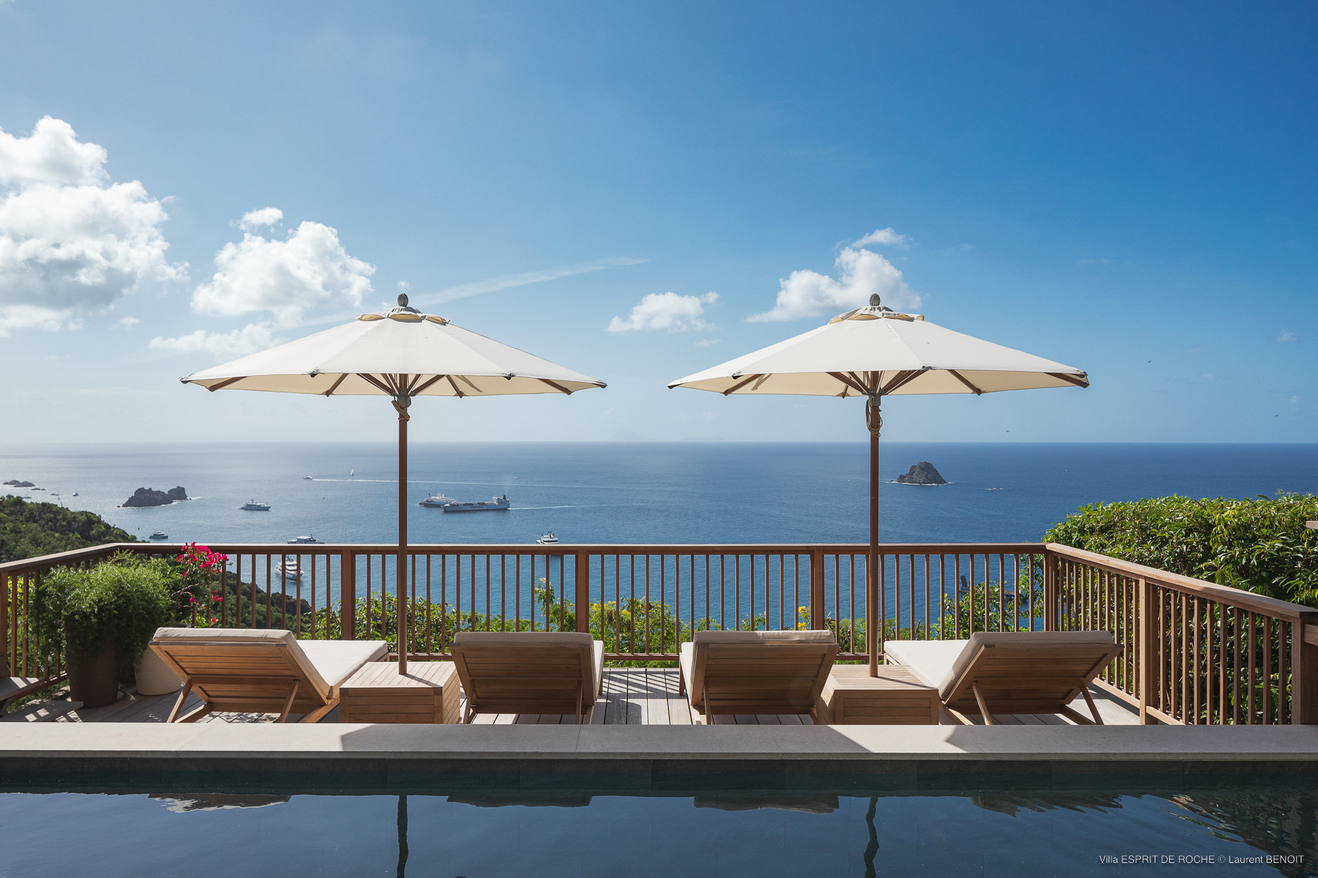 two pairs of wooden sun loungers with large white umbrellas at the edge of an infinity pool, with a stunning blue ocean view beyond