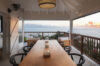 a covered outdoor dining area on a terrace at dusk with a long wooden table, black chairs, and a view of the sunset over the ocean
