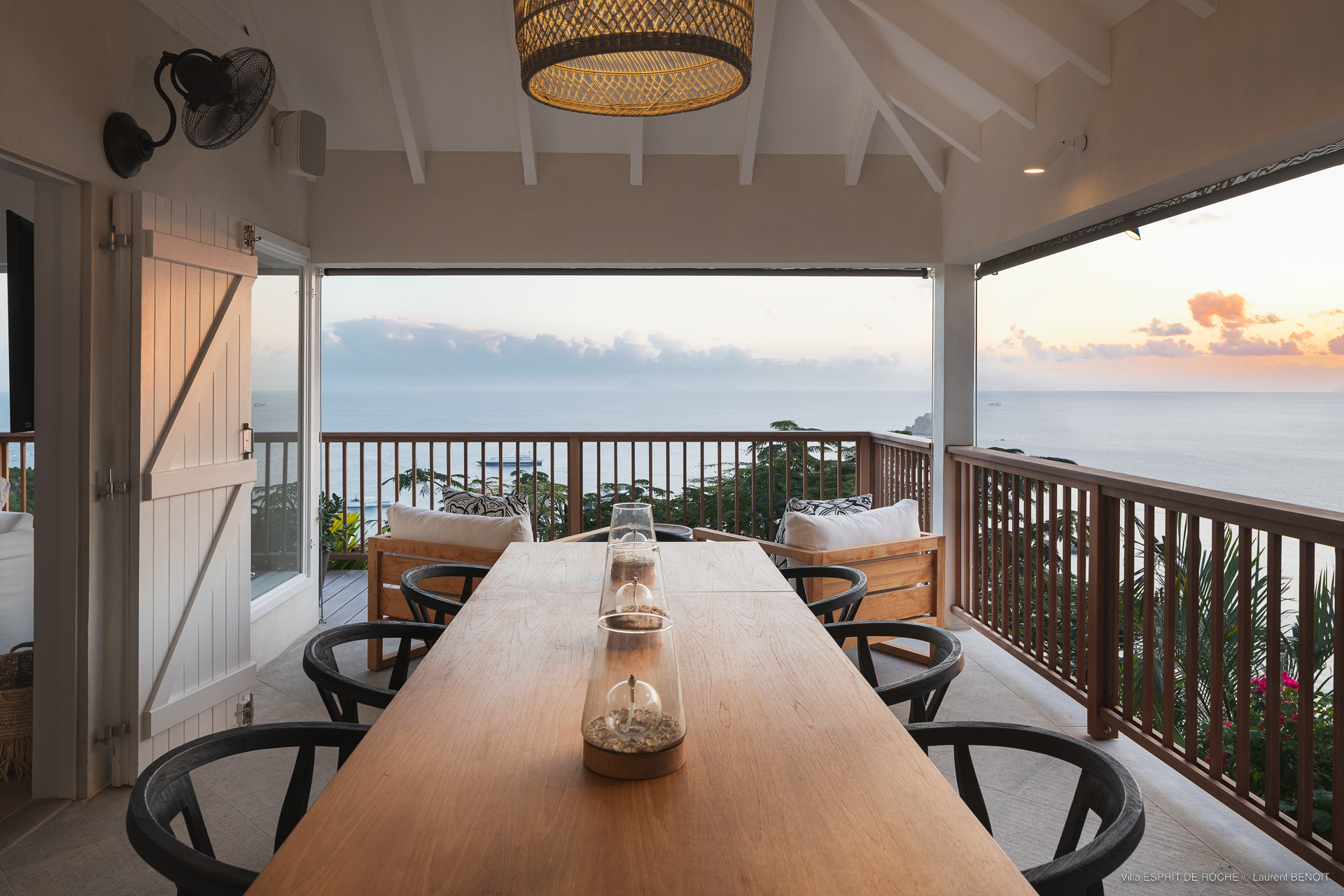 a covered outdoor dining area on a terrace at dusk with a long wooden table, black chairs, and a view of the sunset over the ocean