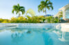 A clear, calm jacuzzi with water reflecting the surrounding palm trees and the bright blue sky. The edge of the villa and distant hills are visible in the background.