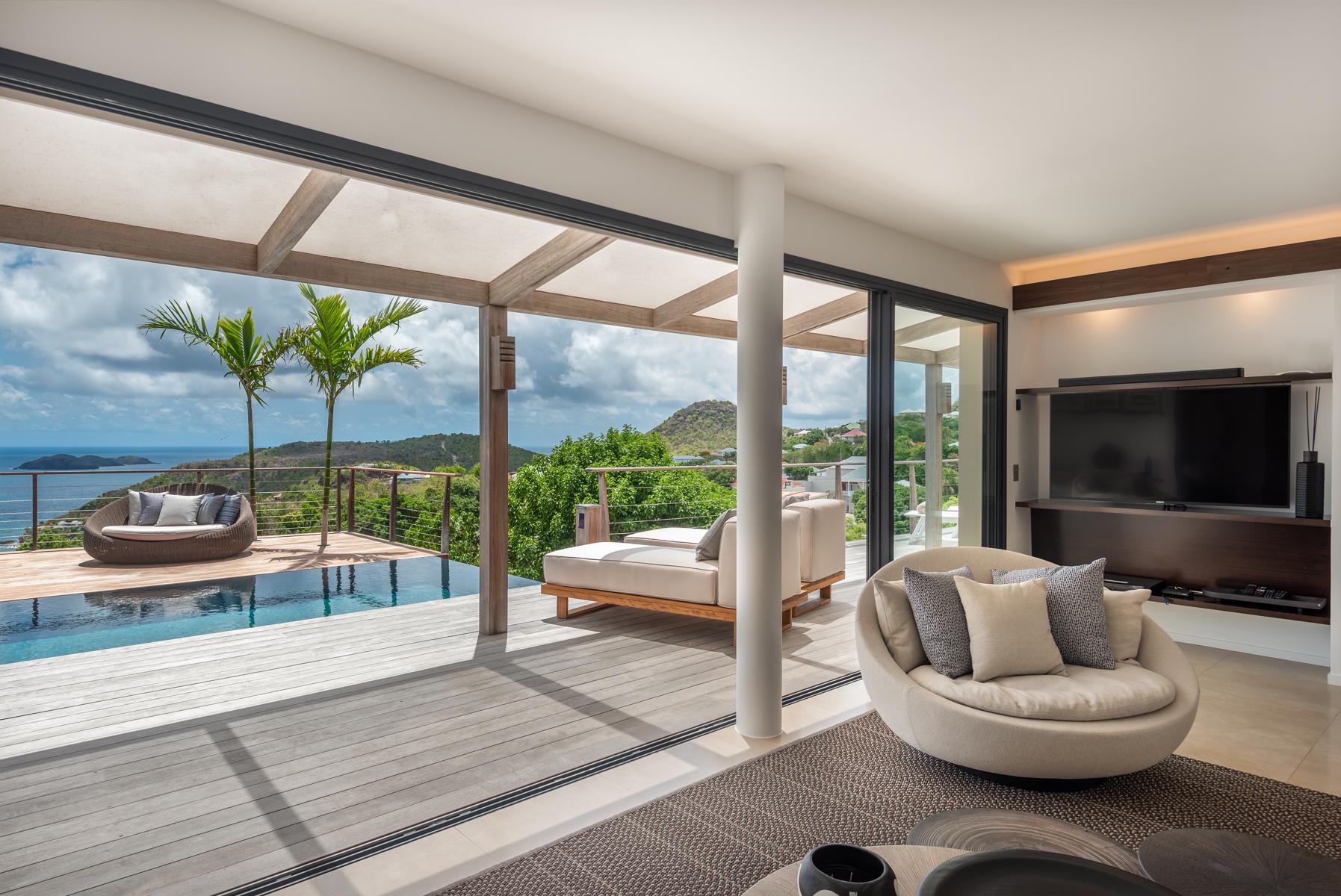 Modern living room in a St Barts villa opening to a pool terrace with sunbeds, palm trees, and panoramic ocean views.