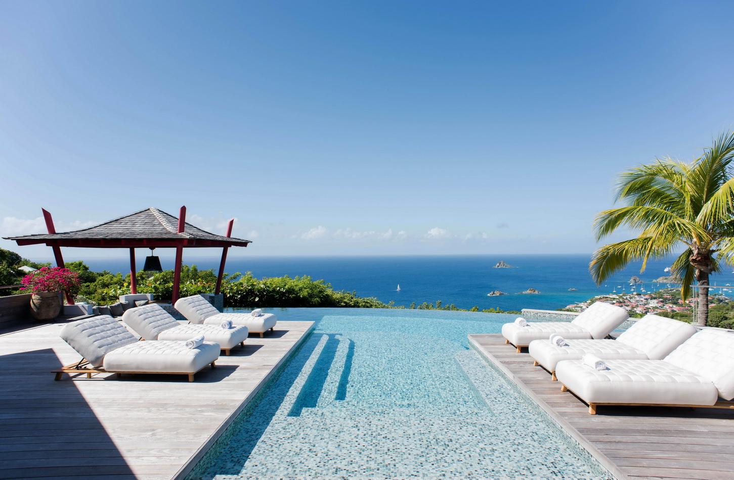 Luxury St Barts villa with sleek infinity pool, sun loungers on wooden deck, and shaded outdoor living spaces surrounded by palm trees.