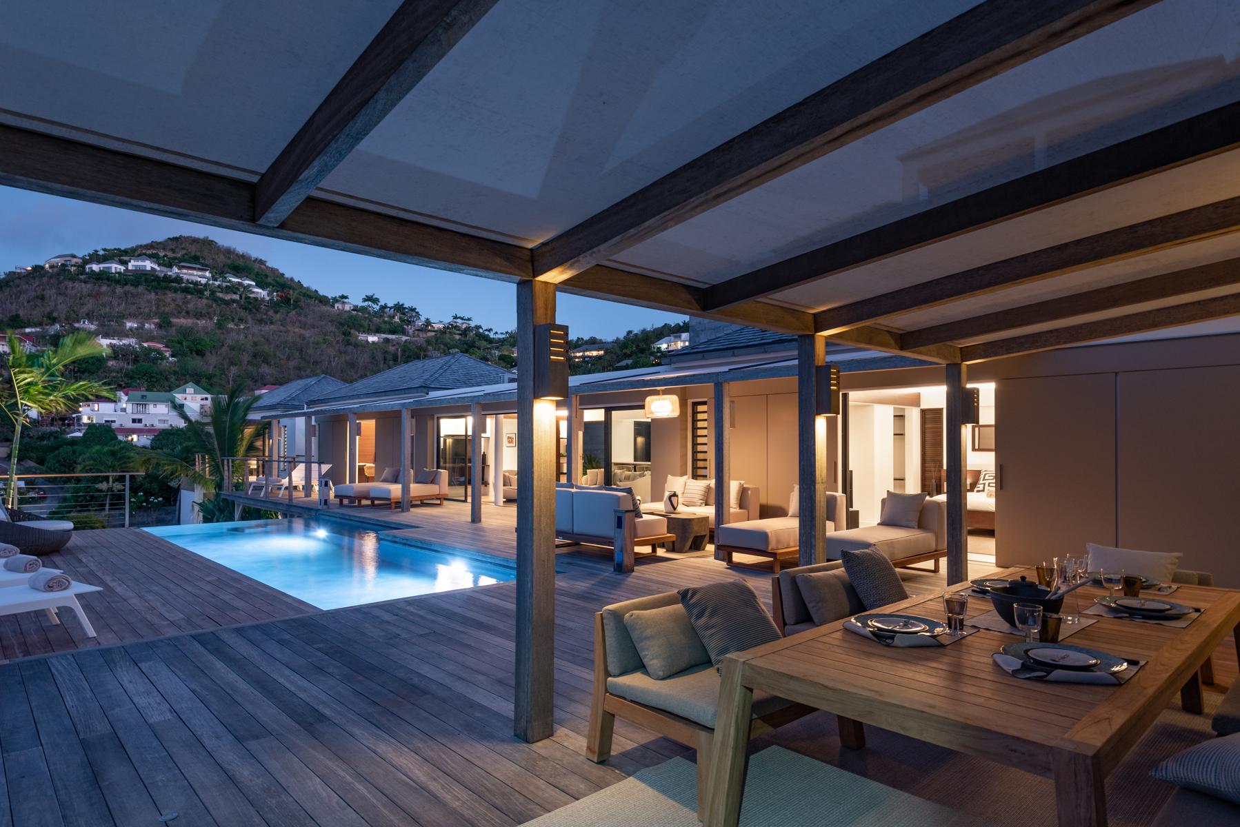 Evening ambiance at a St Barts villa with poolside terrace, alfresco dining table, and warm lighting against hillside views.