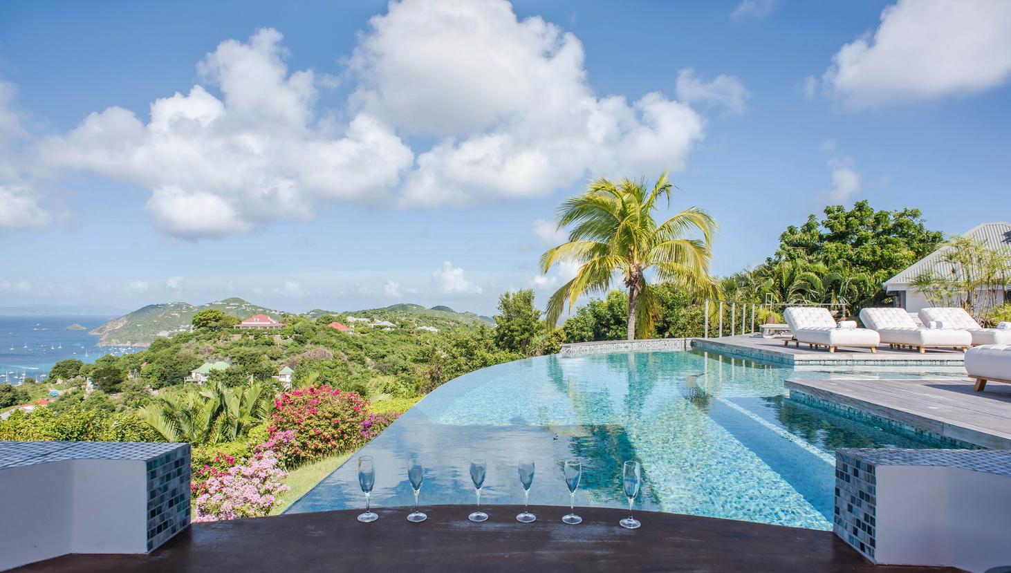 Luxury St Barts villa with sleek infinity pool, sun loungers on wooden deck, and shaded outdoor living spaces surrounded by palm trees.