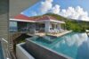 Infinity pool at a St Barts villa with sun loungers on the terrace, modern architecture, and hillside views under blue skies.