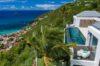 Hillside St Barts villa with infinity pool overlooking palm trees, turquoise sea, and red-roofed coastal village below.