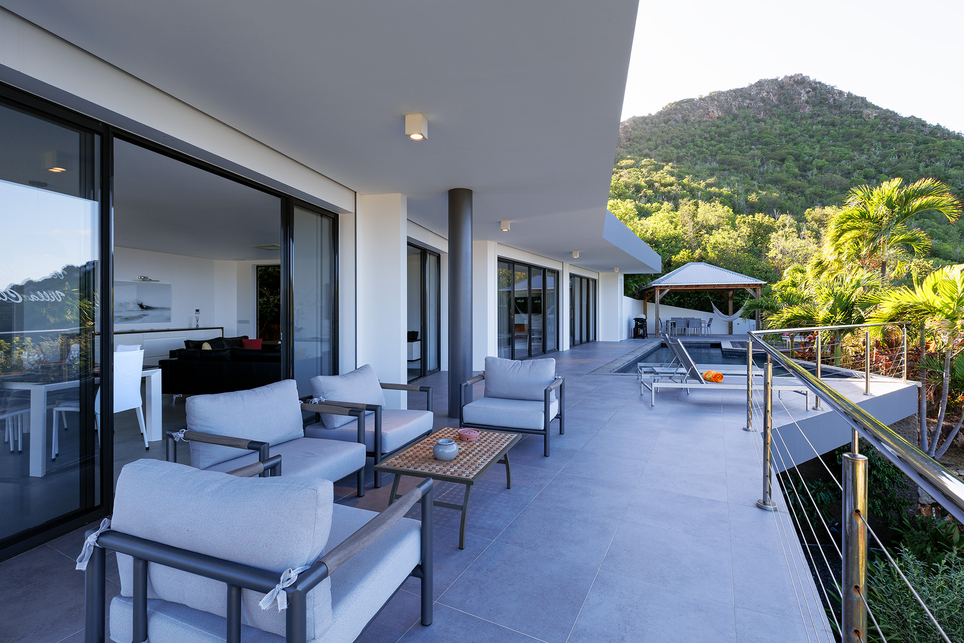 Expansive terrace of a St Barts villa with cushioned lounge chairs, outdoor dining, and views of tropical hillsides.