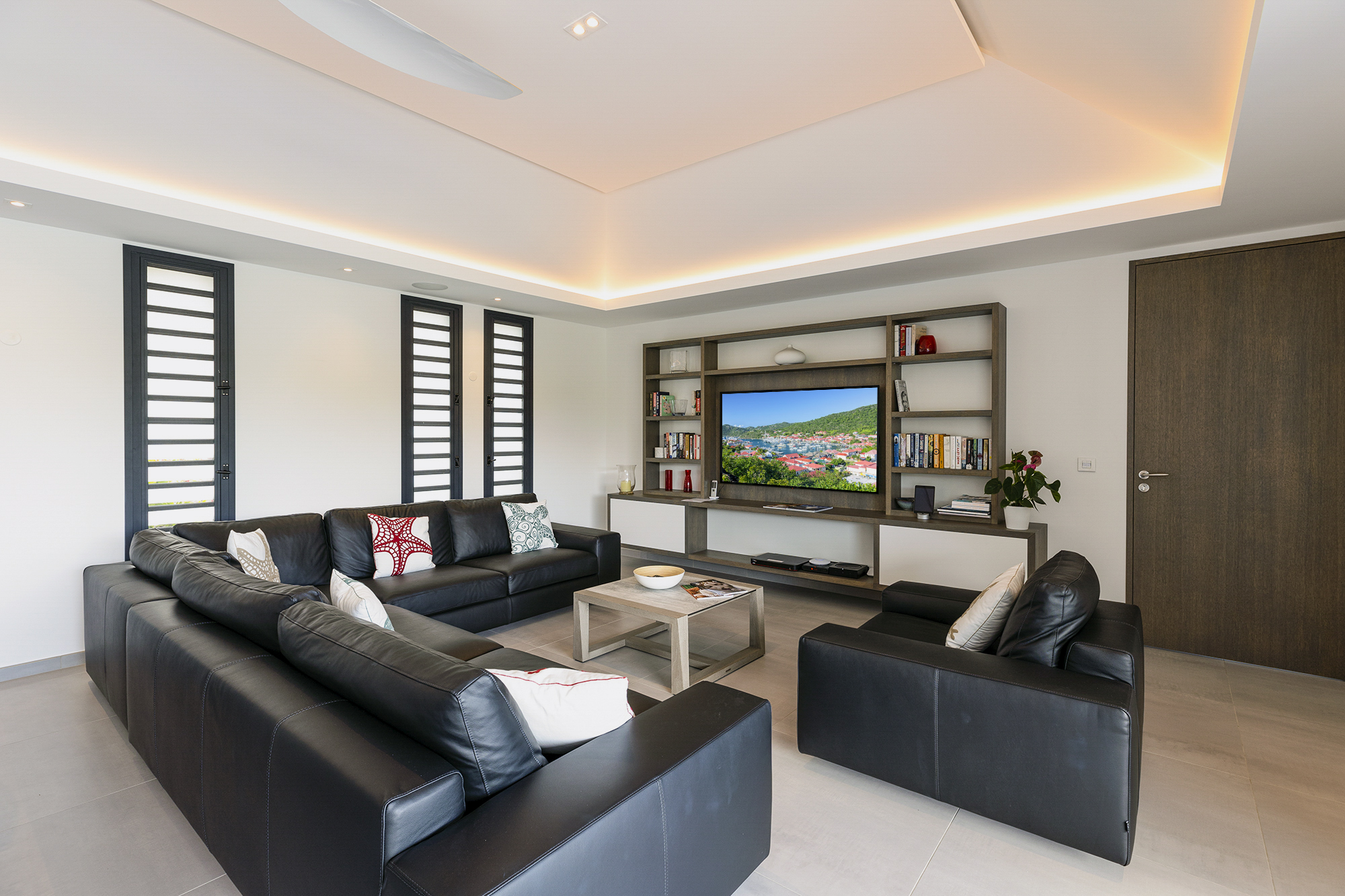 Modern media room in a St Barts villa with black leather sofas, built-in shelving, and large flat-screen TV.