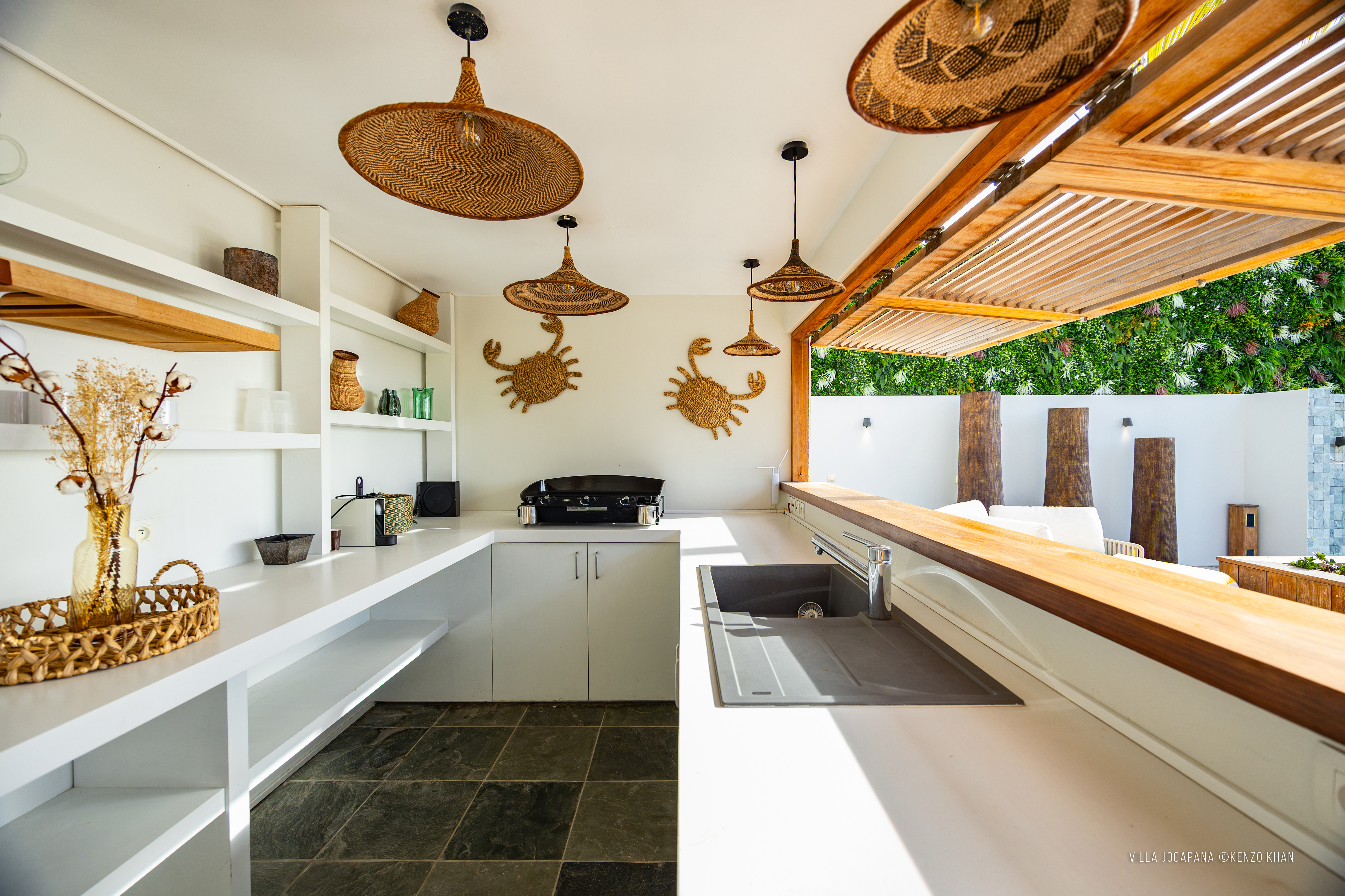 Open-air kitchen at Villa Jocapana featuring woven pendant lights, tropical rattan crab wall décor, sleek countertops, and a bar counter opening onto the terrace.