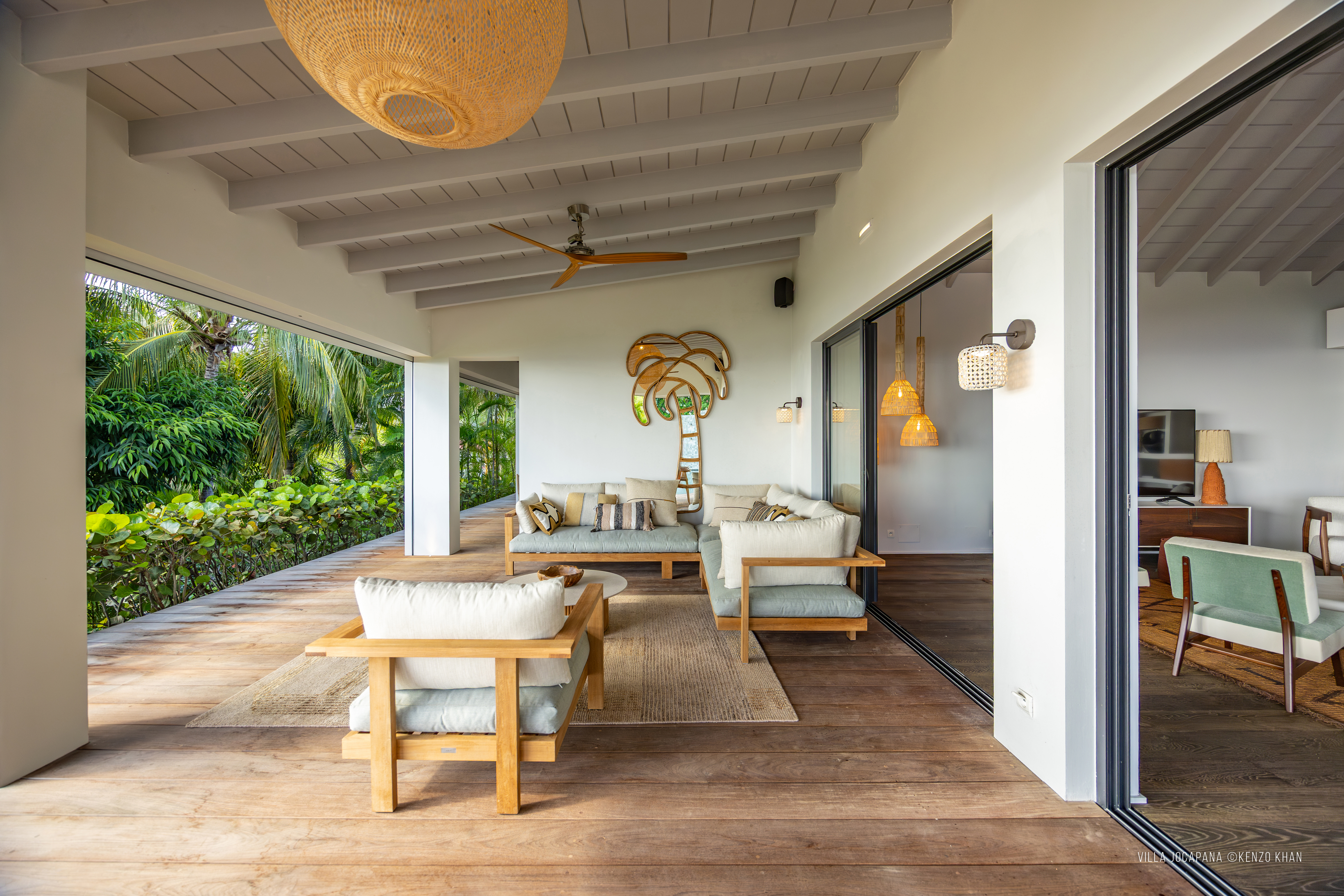 Spacious open living area at Villa Jocapana with natural wood furniture, soft neutral seating, artistic palm tree wall décor, and seamless flow to the indoor lounge and library.