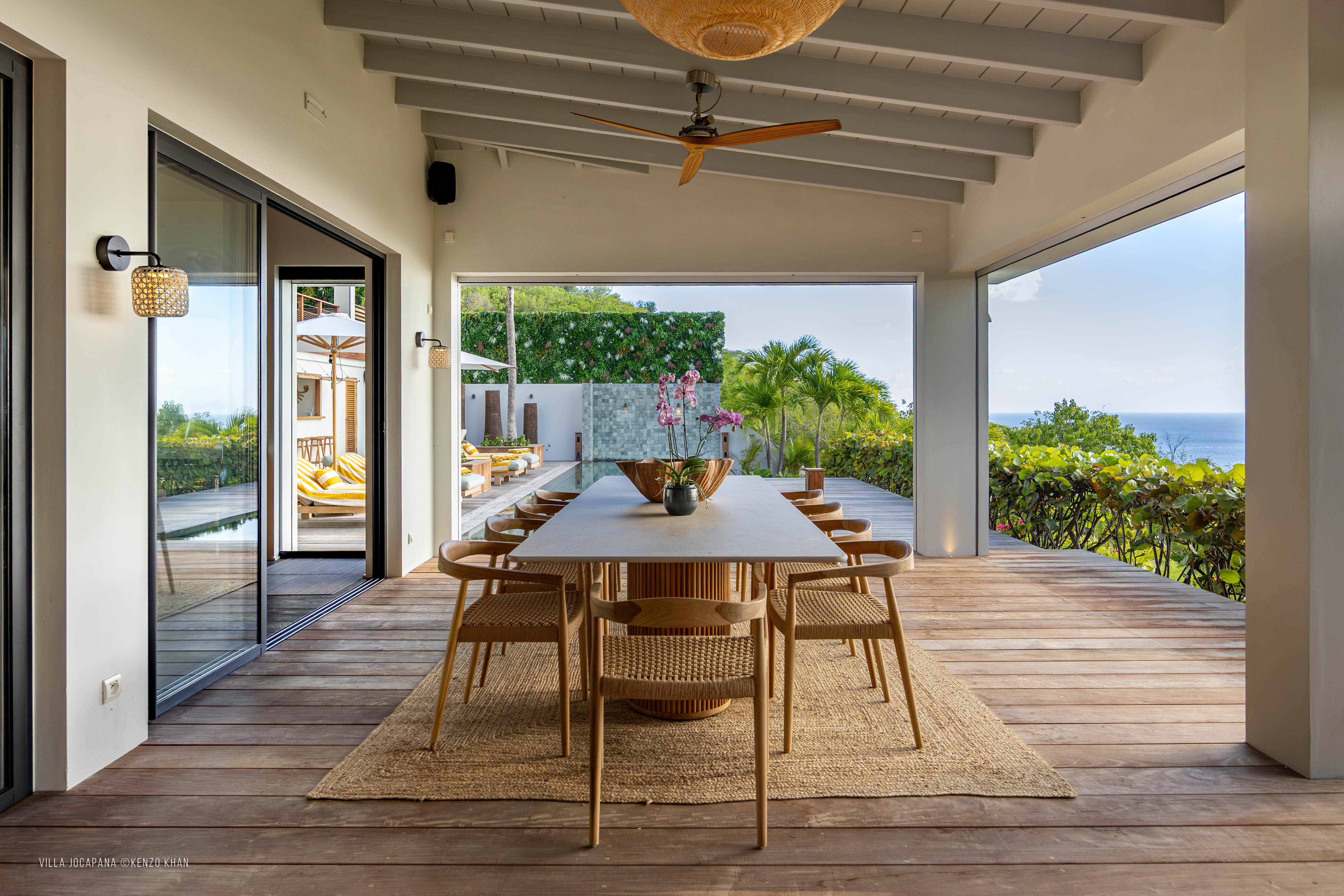Open-air dining terrace at Villa Jocapana with a modern wooden table, woven chairs, orchids as a centerpiece, and panoramic ocean views framed by lush greenery.